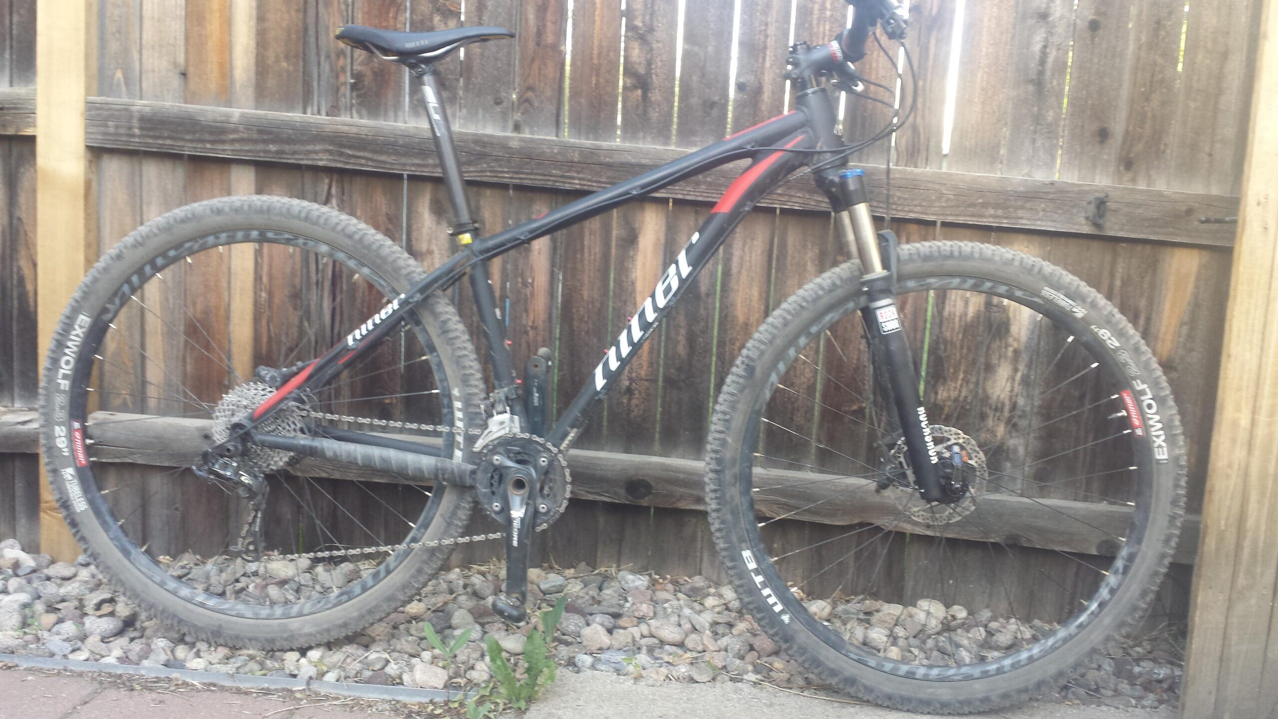 Niner EMD 9: Image of a mountain bike leaning against a wooden fence. The bike features a black and red frame, large tires, and a visible gear system. The surroundings include gravel and some greenery, indicating an outdoor setting.