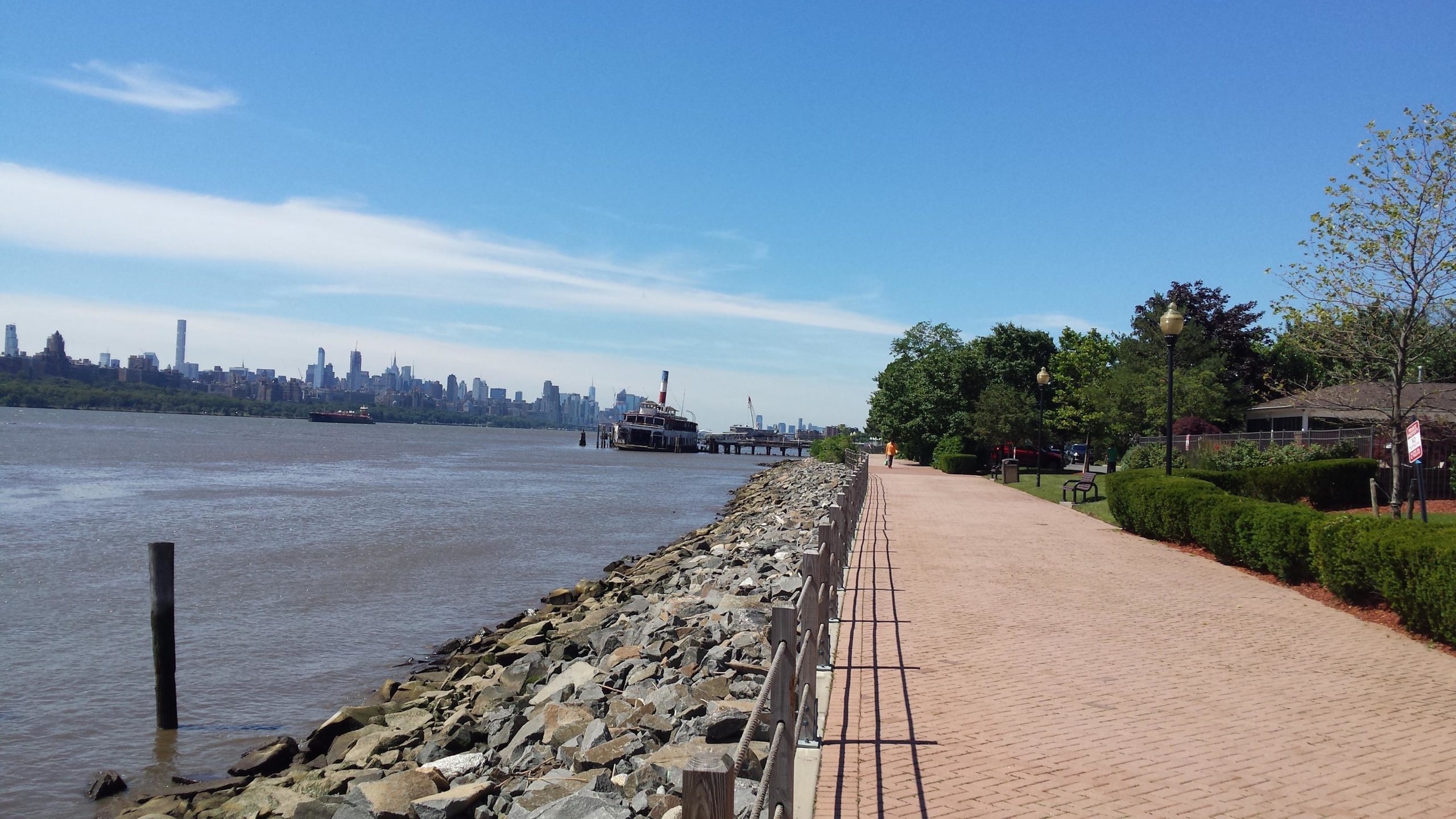 A scenic view of a riverside promenade lined with greenery and benches, featuring a rocky shoreline on one side and a wooden pier with a boat docked on the other. In the background, the skyline of a city is visible under a clear blue sky. West Street Greenway mountain bike trail.