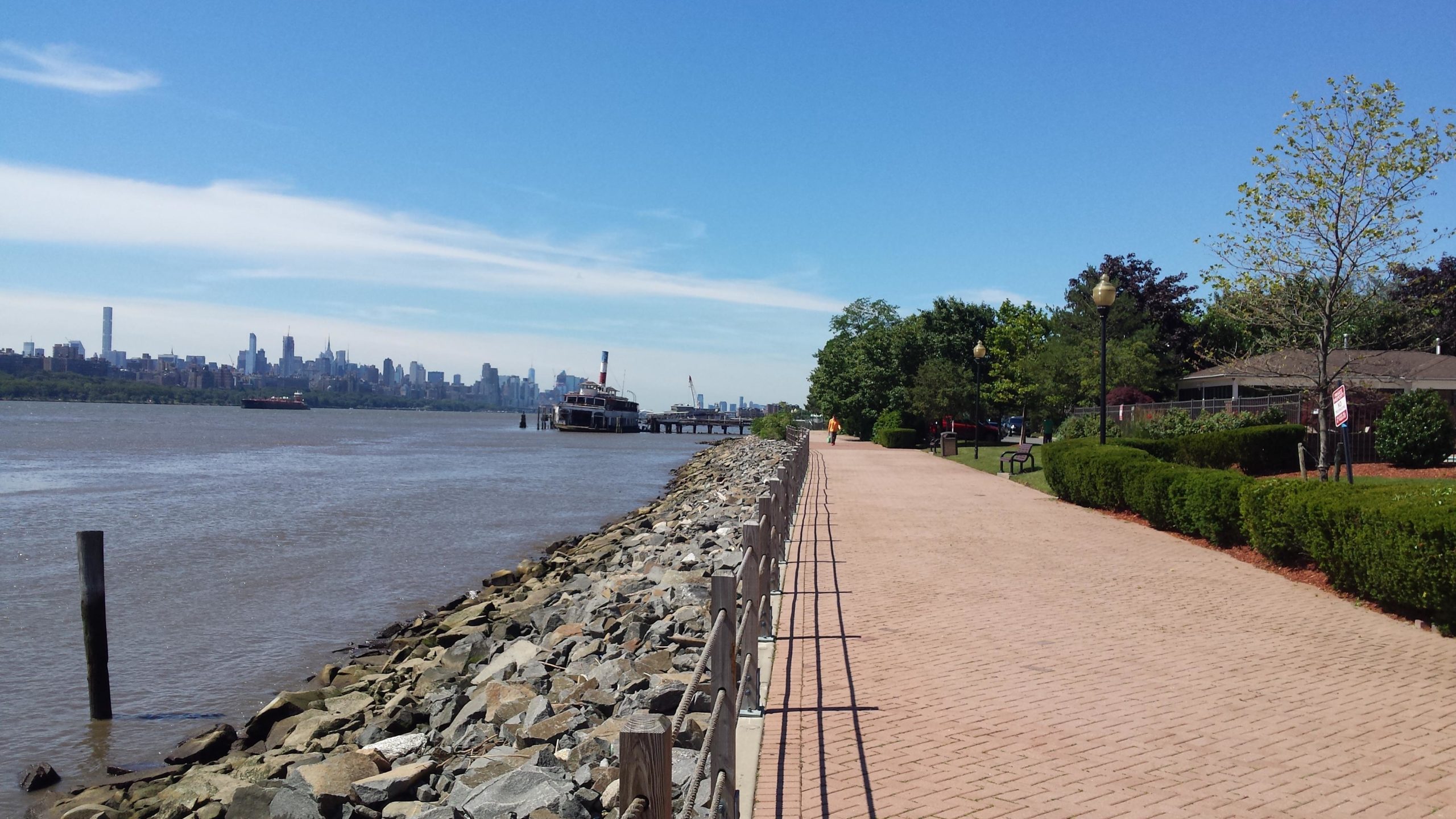 A scenic view along a waterfront walkway featuring a paved path lined with greenery and benches. In the background, a river with a boat is visible, and the skyline of a city can be seen across the water under a clear blue sky. West Street Greenway mountain bike trail.