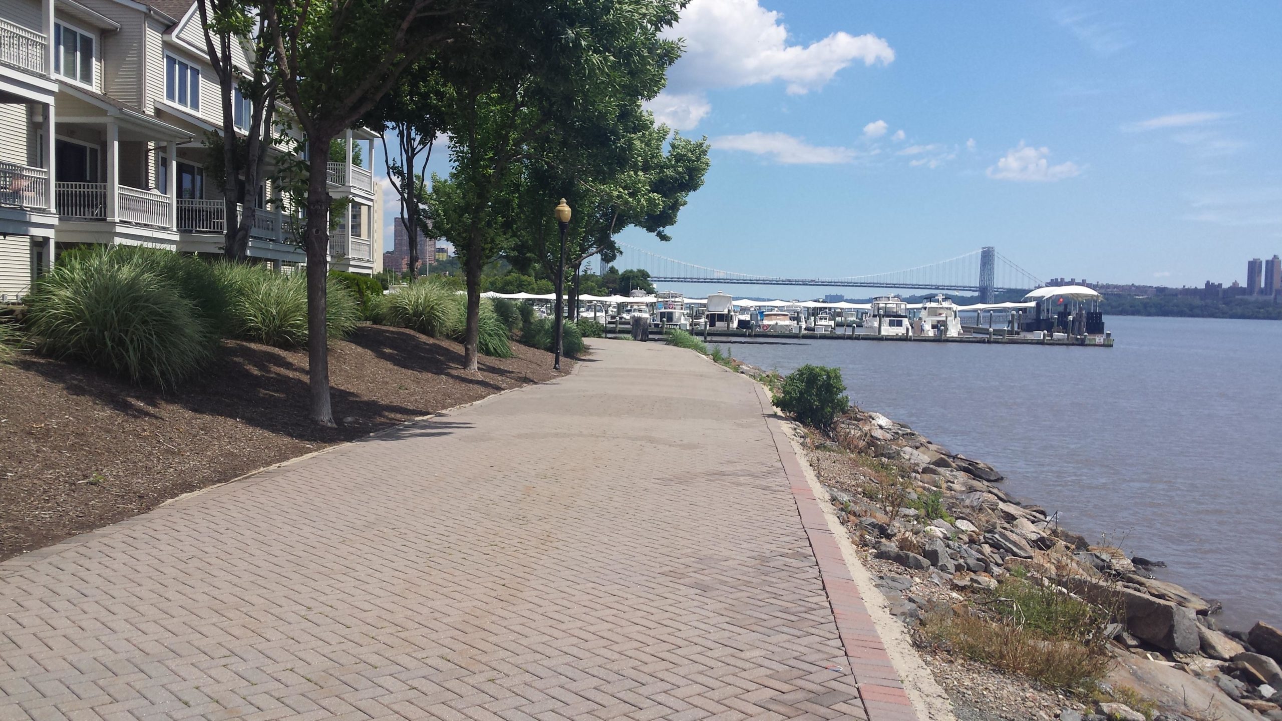 Pathway along a river with residential buildings on one side, grassy areas, and a marina with boats on the water. A bridge is visible in the background under a blue sky with clouds. West Street Greenway mountain bike trail.