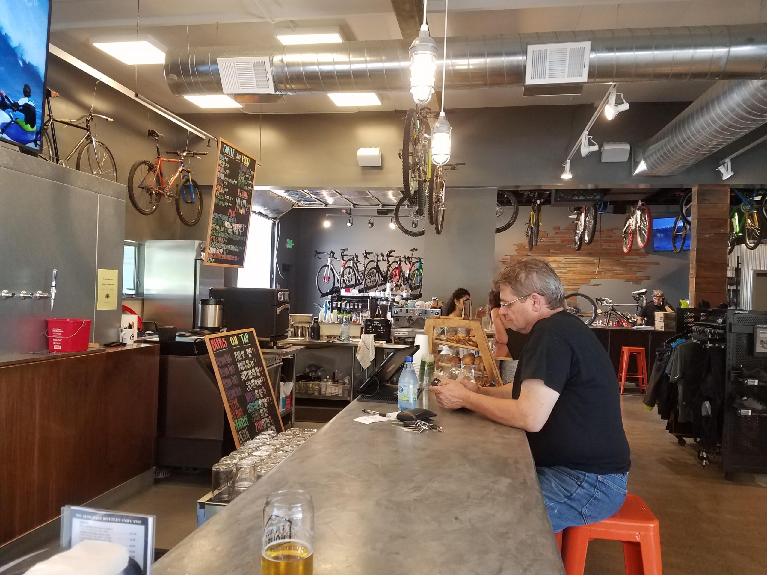 A cozy café and bike shop interior featuring a man sitting at the counter, looking at a menu. The space is adorned with colorful bicycles hanging from the ceiling and walls, a chalkboard displaying food and drink options, and a bar with taps. In the background, several customers are enjoying the atmosphere.