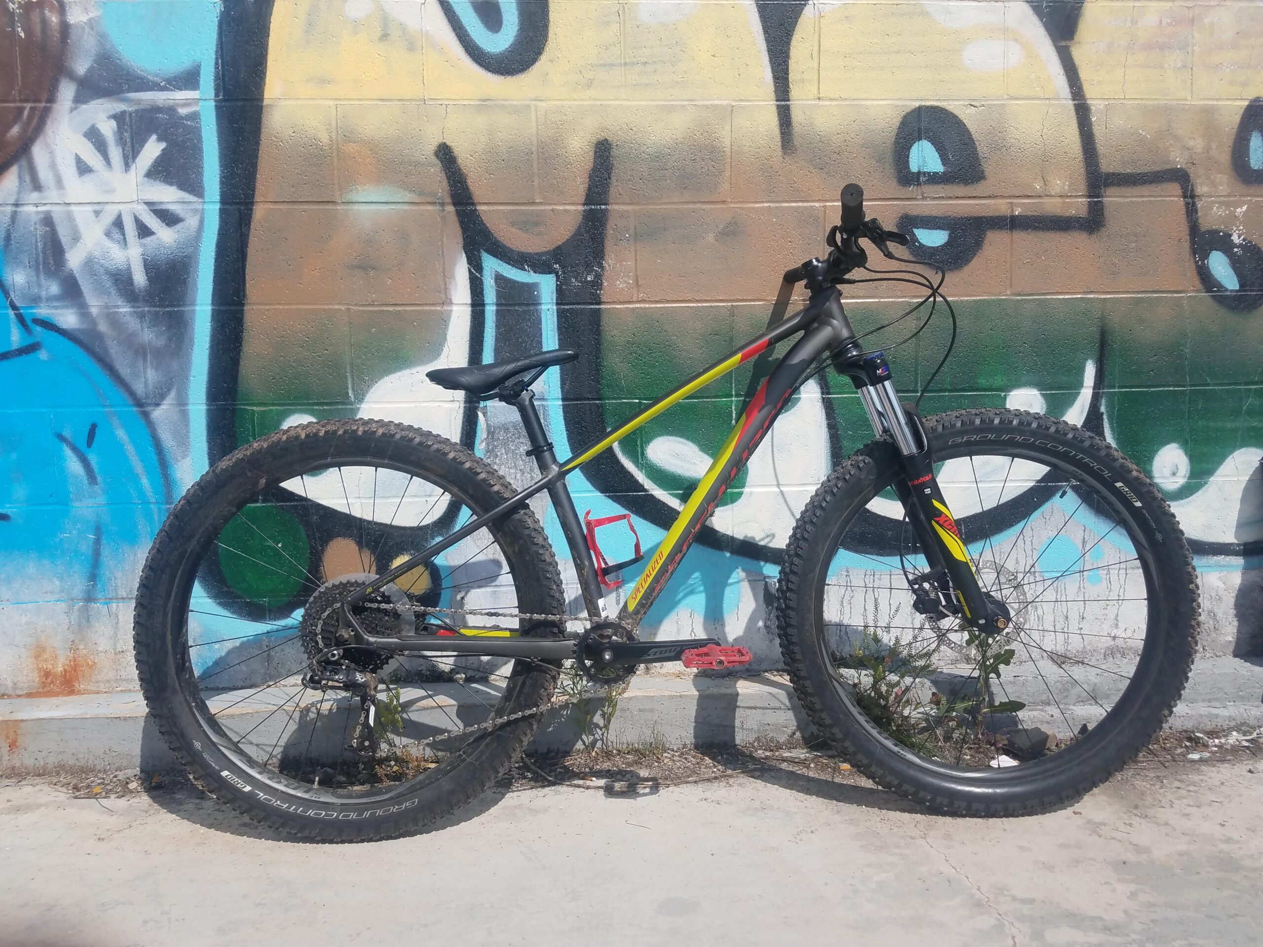 Specialized Fuse: A black and yellow mountain bike with thick tires, leaning against a colorful graffiti wall. The wall features blue and brown hues, with artistic designs in the background. Sparse weeds grow near the bike, and the sun casts shadows on the concrete surface.