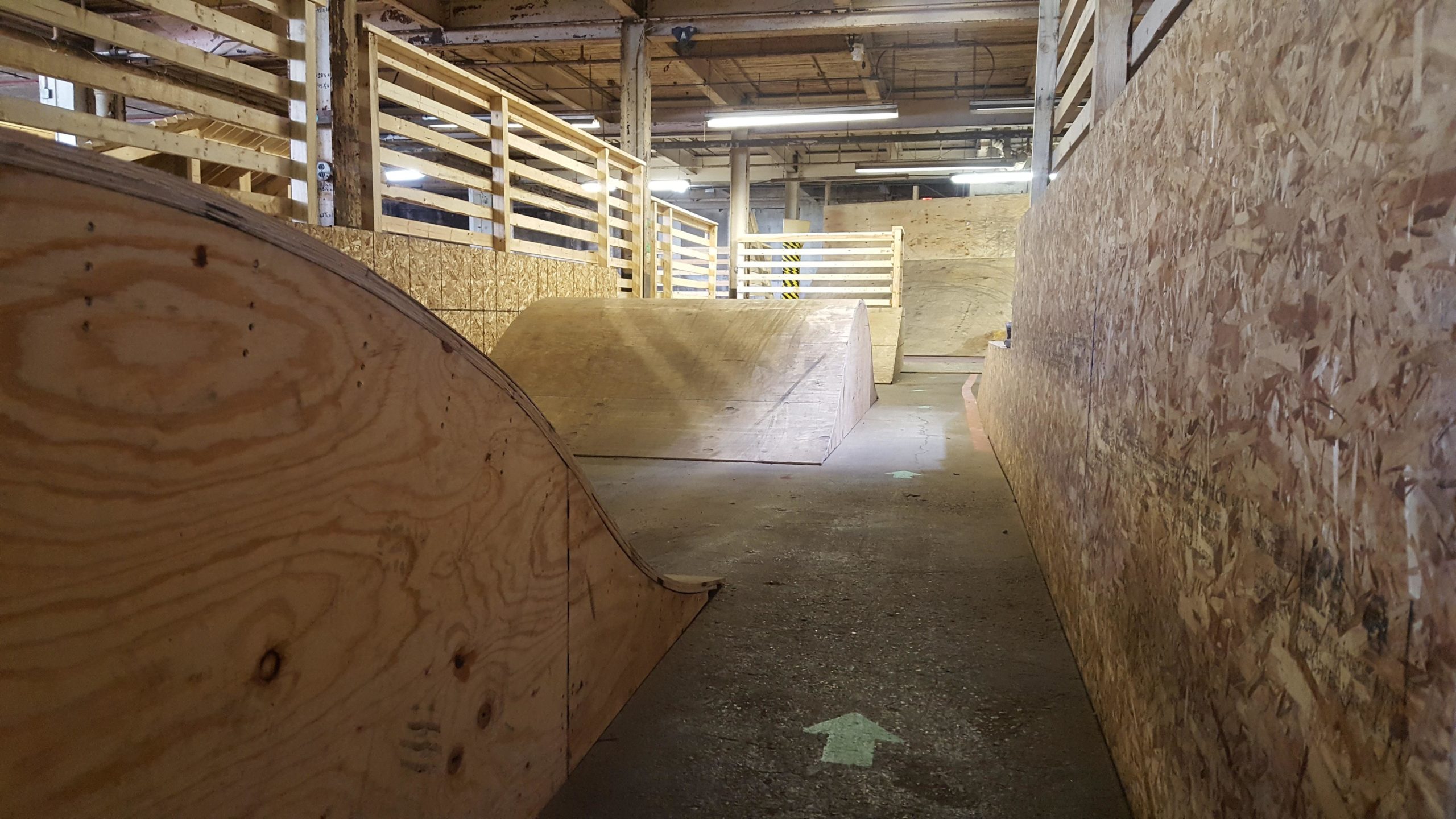 A wooden bike or skateboarding park featuring various ramps and obstacles, viewed from the ground level. The space is well-lit with overhead lights and has a wooden railing along the sides. 

 Mike's Indoor Bike Park mountain bike trail.
