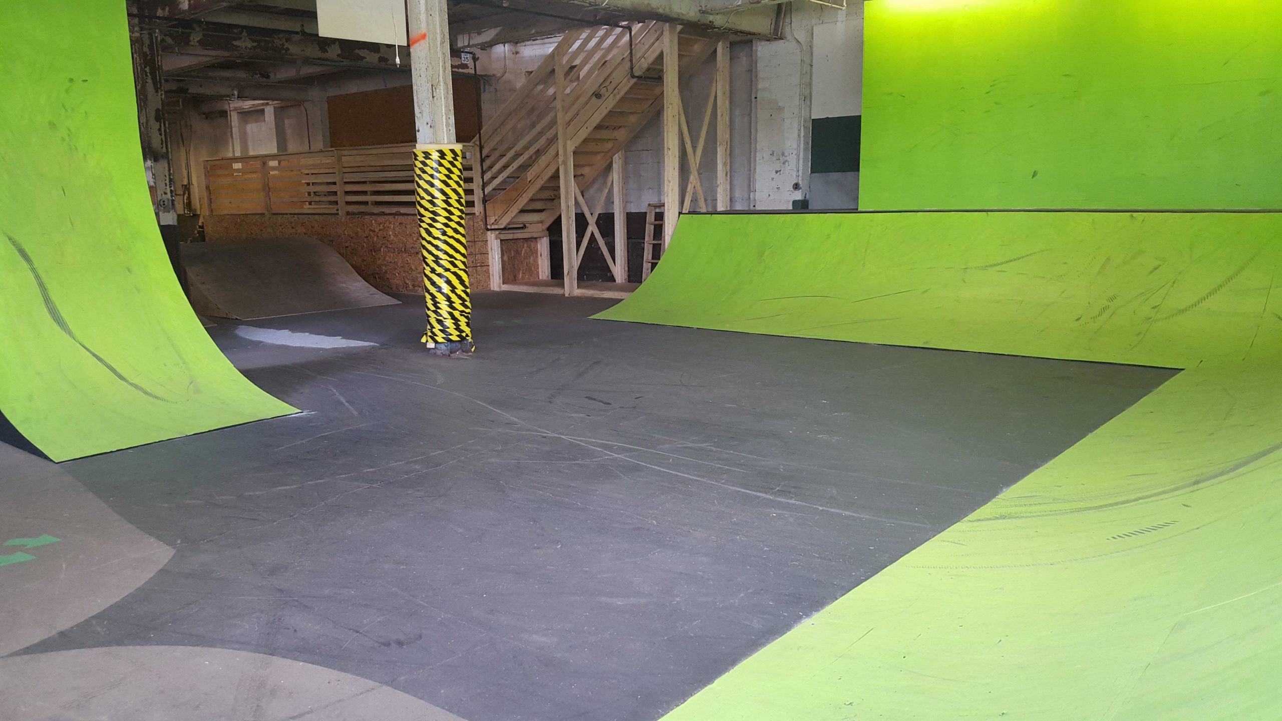 Indoor skate park featuring bright green ramps and a black floor, with a wooden staircase and safety-striped pillar visible in the background. Mike's Indoor Bike Park mountain bike trail.