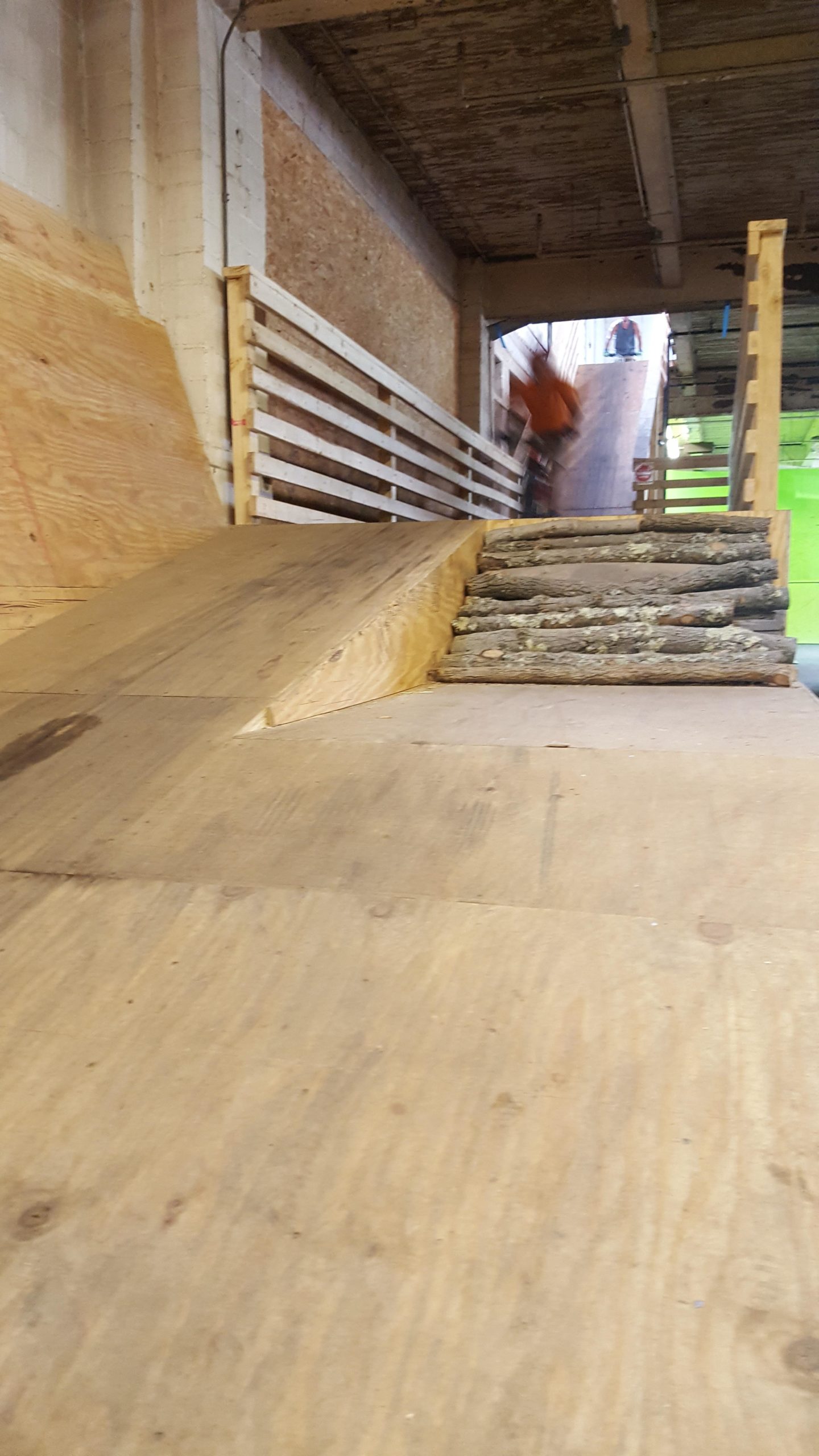 A cyclist performing a trick on a wooden ramp, which leads to a series of logs arranged as obstacles. The indoor setting features a partially visible background where other individuals can be seen observing. The ramp is made of plywood, and the environment has a rustic feel with exposed building materials. Mike's Indoor Bike Park mountain bike trail.