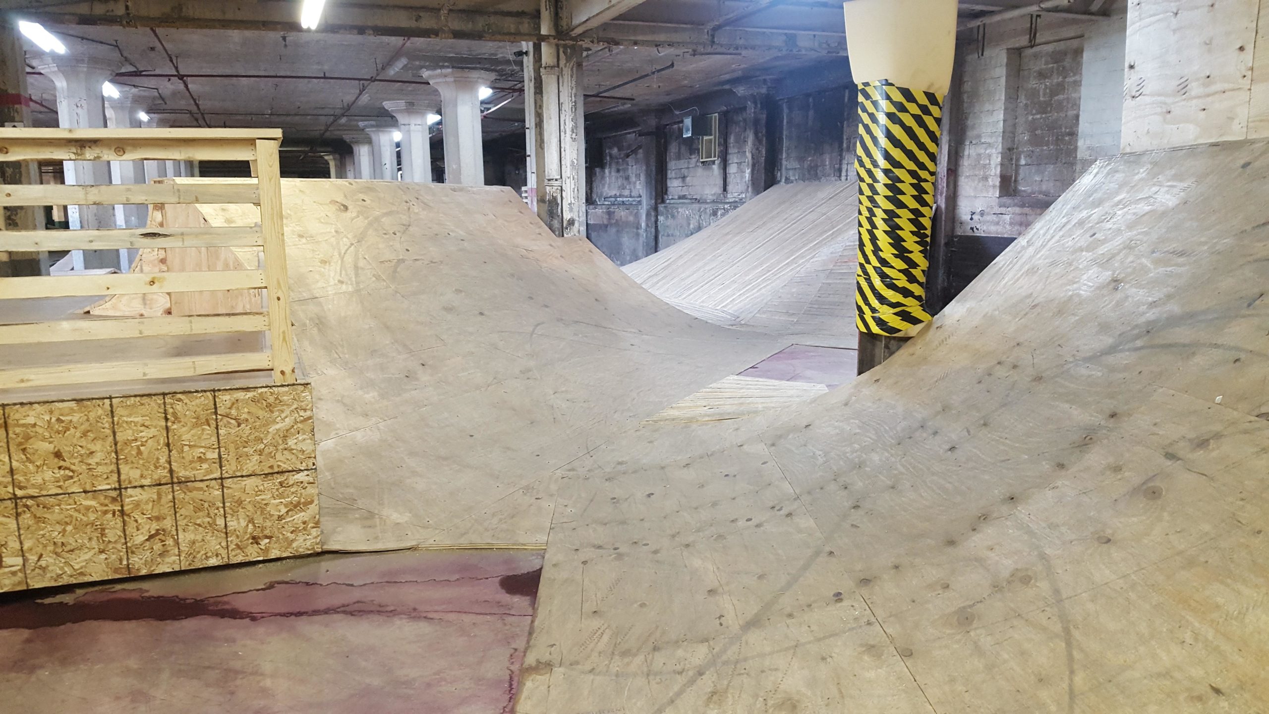 A spacious indoor skatepark featuring wooden ramps and jumps. The facility has a high ceiling with exposed beams and columns, and the floor is a mix of concrete and painted surfaces. One corner features a wooden structure with a railing, and there are caution-striped poles for safety. The atmosphere suggests a recreational space for skateboarding or BMX. Mike's Indoor Bike Park mountain bike trail.