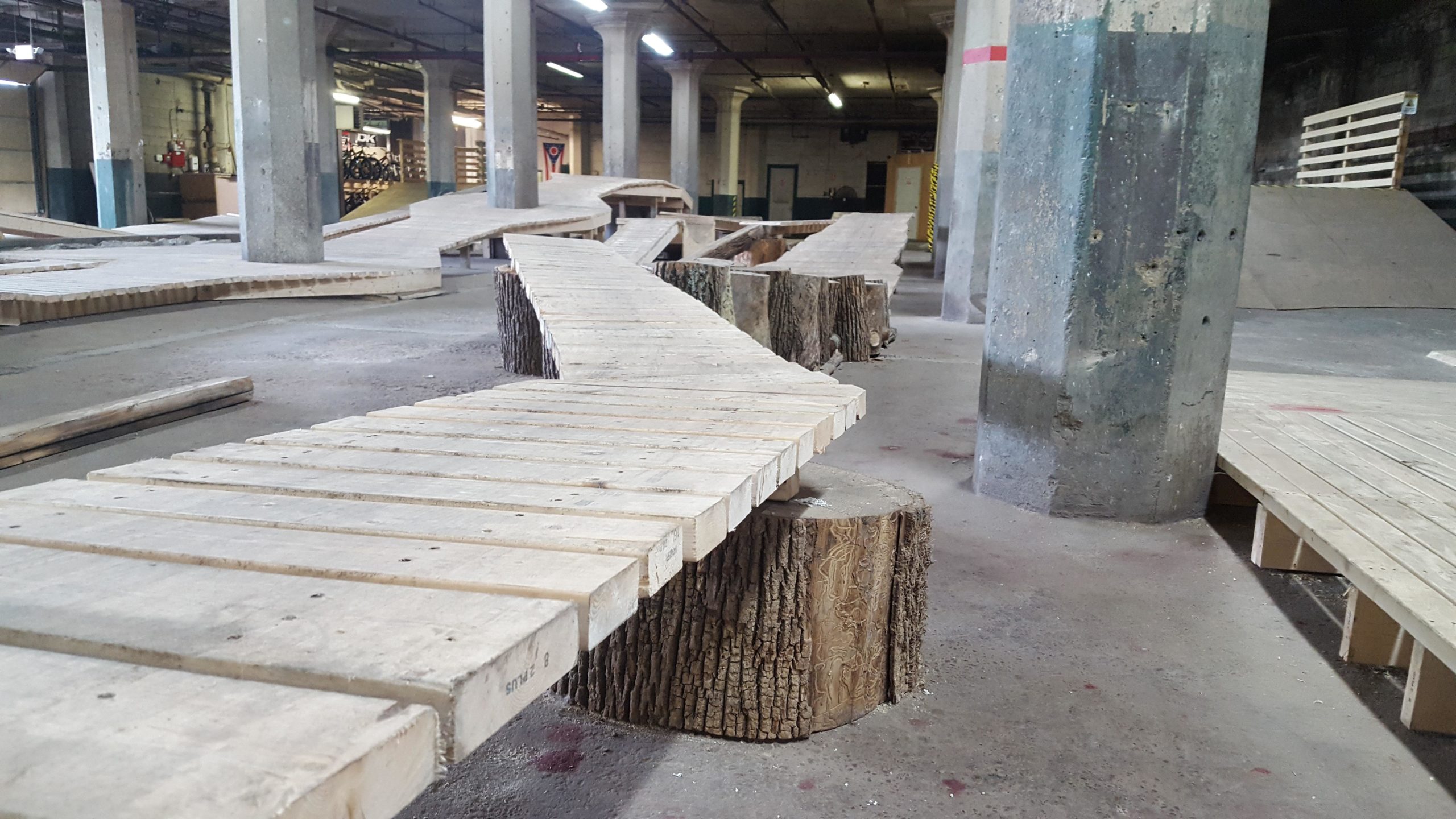 Indoor biking or skateboarding park featuring wooden ramps and platforms made from boards and tree logs, set in a spacious industrial environment with exposed concrete pillars and dim lighting. Mike's Indoor Bike Park mountain bike trail.