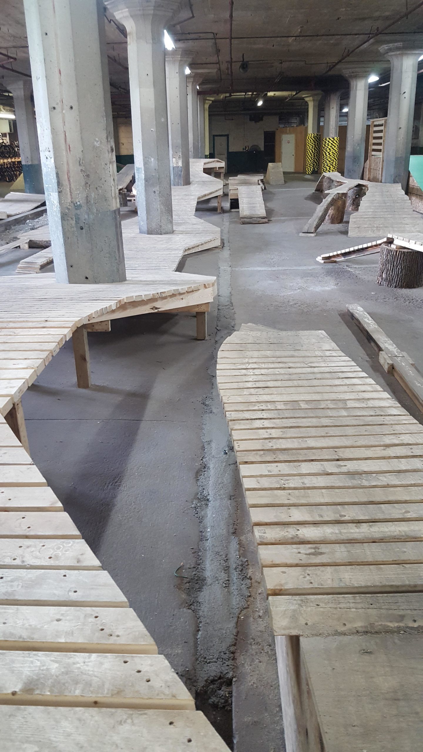 Indoor space featuring a network of wooden track-like structures made from planks, arranged in a winding pattern across a concrete floor. The area is well-lit, with exposed concrete pillars and a slight industrial look, suggesting it may be used for activities like biking, skating, or creative play. Some natural wood elements are visible, providing a contrast to the man-made surfaces. Mike's Indoor Bike Park mountain bike trail.