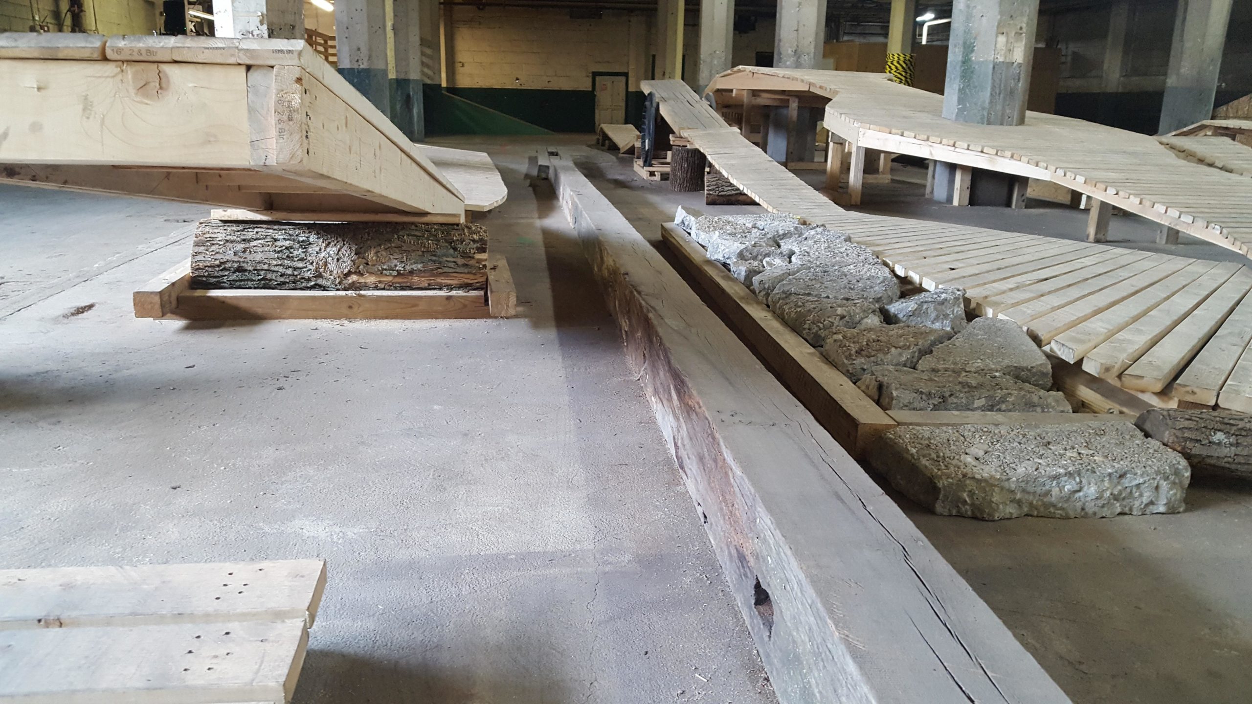 A spacious indoor area featuring various wooden structures and surfaces, including ramps and platforms made from planks and logs. Concrete and wooden materials are arranged on the ground, with a focus on textures and construction elements. The environment appears to be a workshop or design space. Mike's Indoor Bike Park mountain bike trail.
