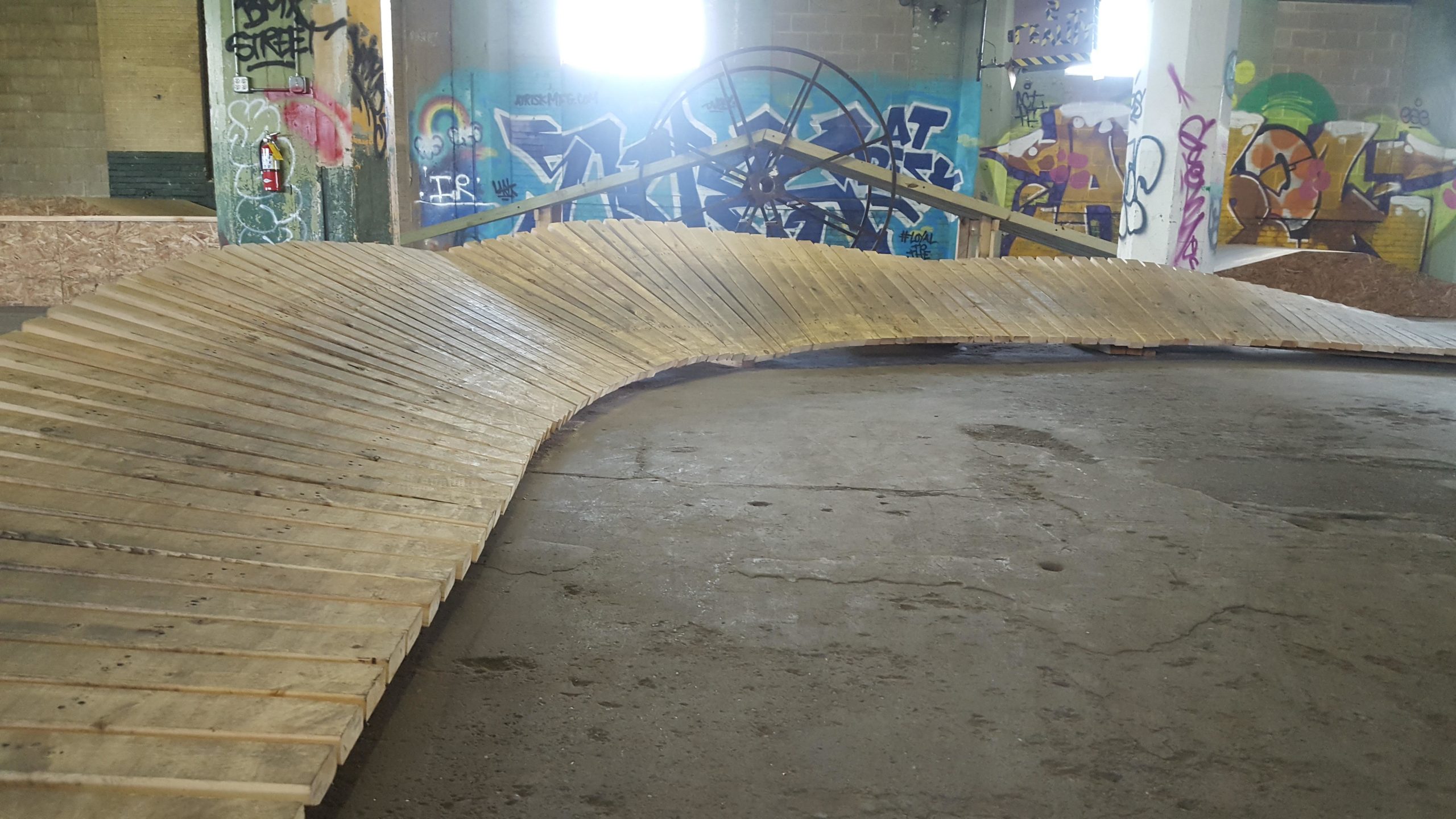 A wooden ramp or half-pipe structure designed for extreme sports, set in an industrial space with colorful graffiti on the walls. The ramp curves along the ground, showcasing its slatted wooden surface, while a large wheel is visible in the background against the vibrant wall art. Mike's Indoor Bike Park mountain bike trail.