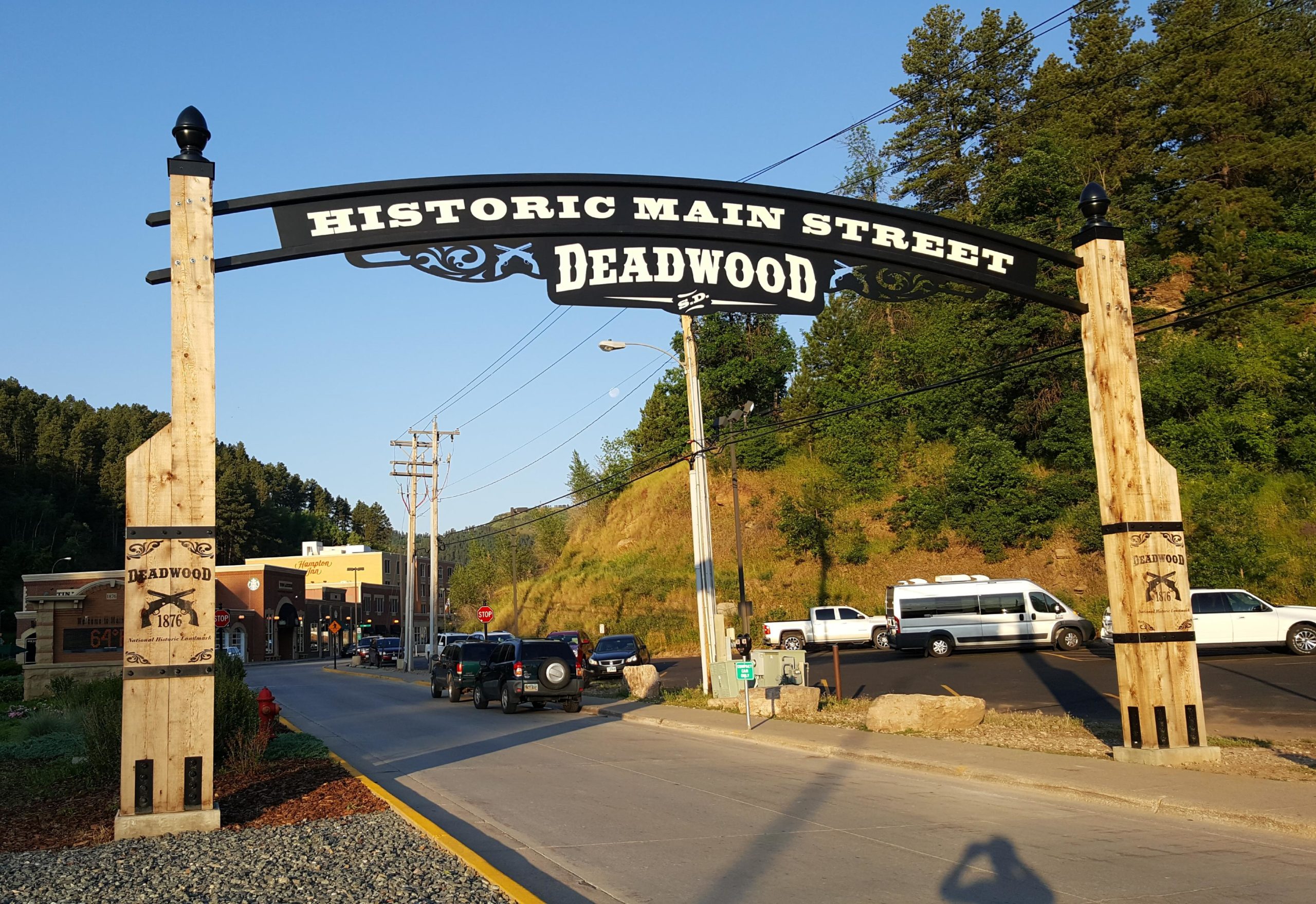 "Sign at the entrance of Historic Main Street in Deadwood, South Dakota, featuring wooden posts and a decorative black arch. The street is visible with parked cars, surrounded by hills and greenery under a clear blue sky." The George S. Mickelson Trail mountain bike trail.
