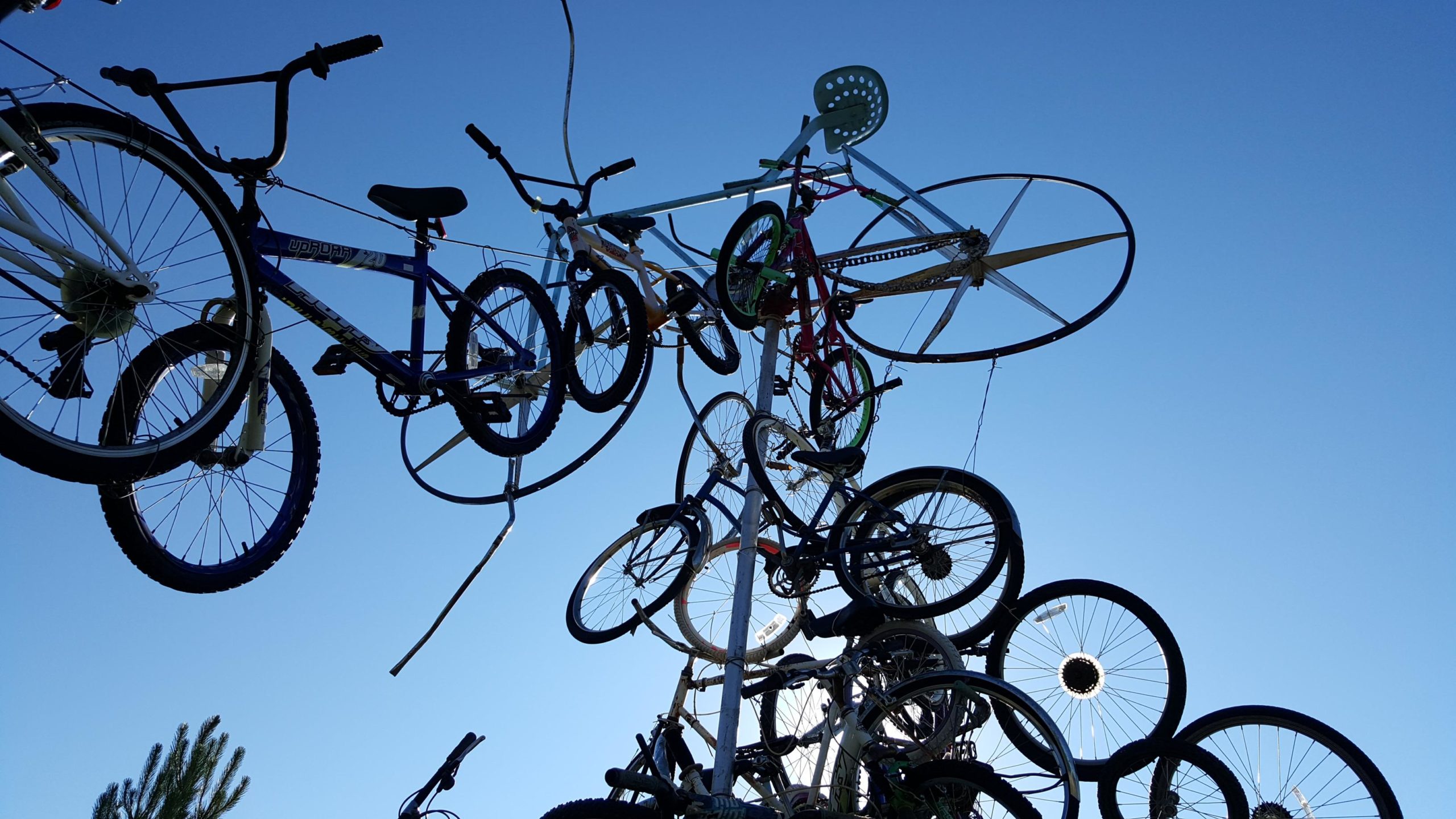 A cluster of bicycles suspended in the air, showcasing a variety of bike frames and wheels against a clear blue sky. The arrangement creates an artistic and playful visual of bicycles intertwined and stacked together, highlighting the different shapes and colors of the bikes. The George S. Mickelson Trail mountain bike trail.