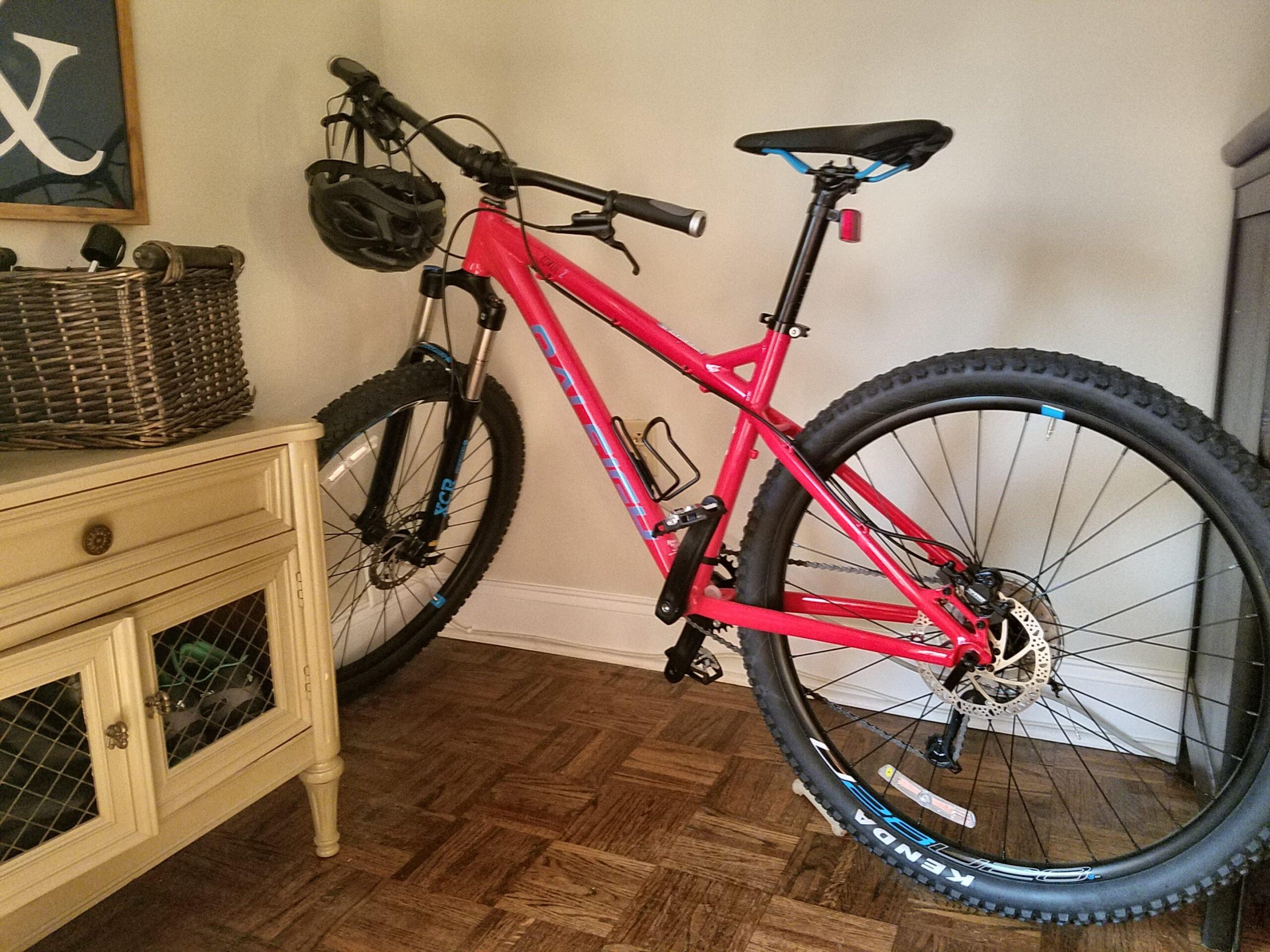 Raleigh Tokul 2: A bright red mountain bike with thick tires stands against a wall indoors. A black helmet is hanging from the handlebars. Next to the bike, there is a woven basket on a light-colored cabinet, and the floor features a wooden parquet pattern.