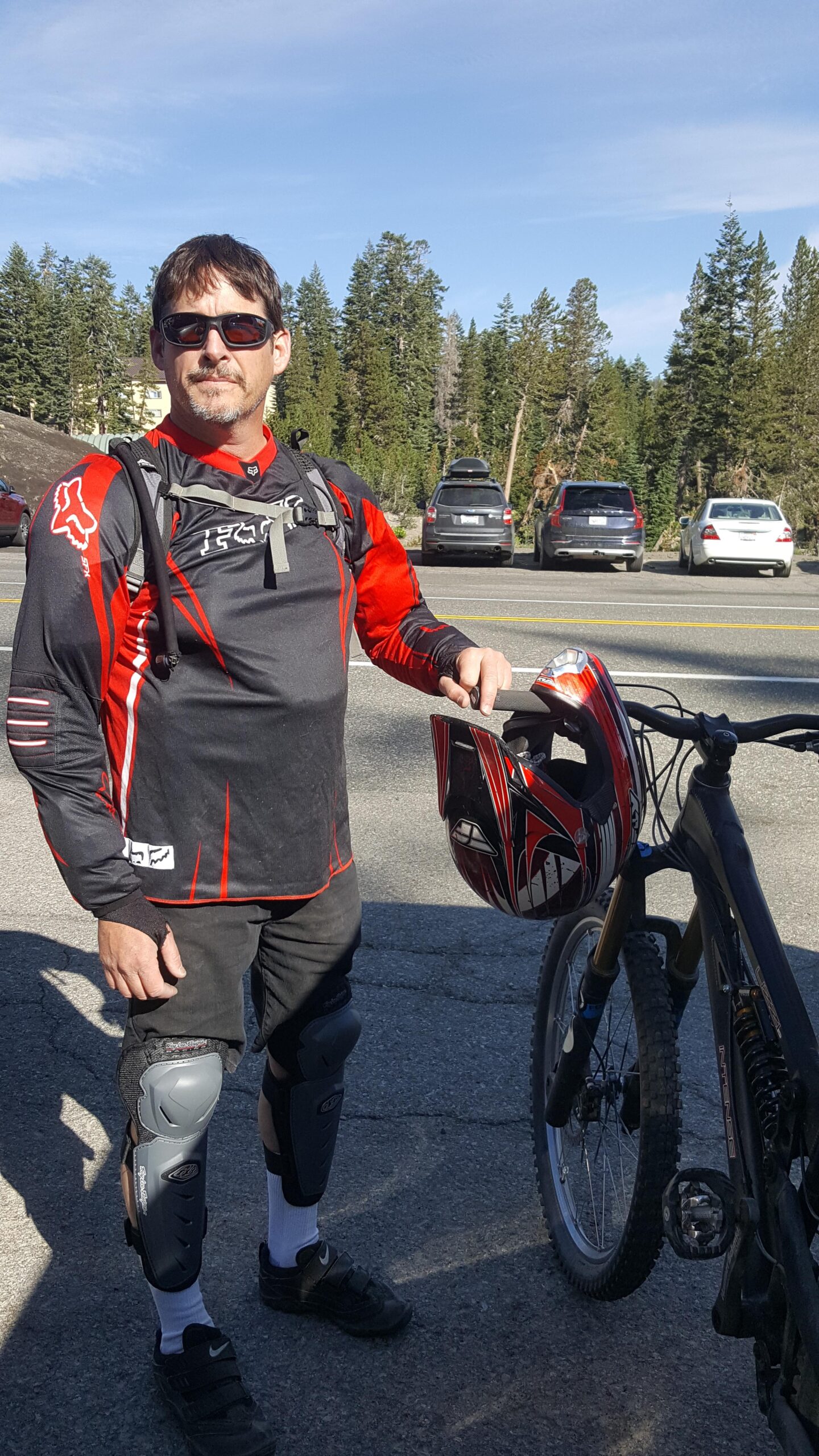 Intense Uzzi vp: A man wearing protective gear, including knee pads and sunglasses, stands next to a mountain bike while holding a helmet. He is dressed in a black and red long-sleeve jersey with a logo, in a parking area surrounded by trees and parked cars.