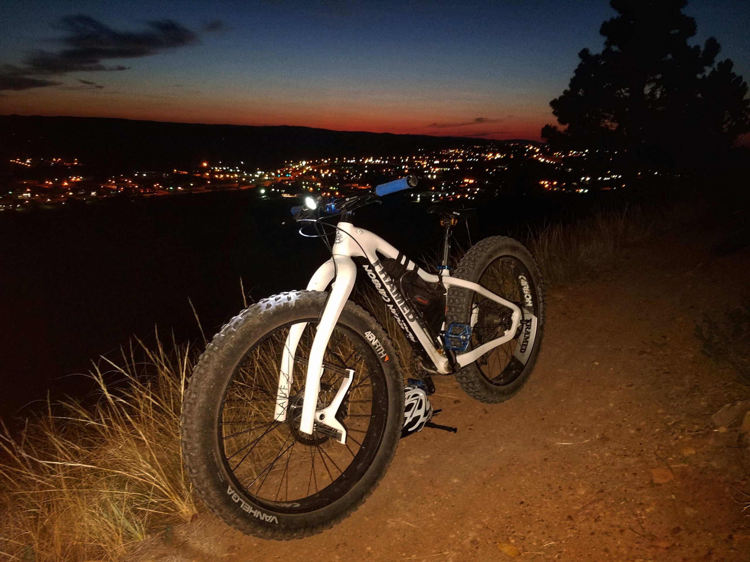 A fat tire bike parked on a dirt path at dusk, overlooking a brightly lit city in the valley below. The sky transitions from deep blue to orange as the sun sets, with silhouettes of trees in the background. HLMP mountain bike trail.