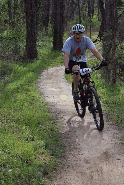 A cyclist wearing a helmet and a gray t-shirt with a logo rides on a dirt trail surrounded by trees and greenery. The rider has a number tag visible on their jersey as they navigate the bend in the path. Lawrence Riverfront Trails mountain bike trail.