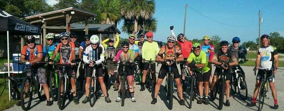 A large group of cyclists, both men and women, poses together on a sunny day in a rural setting. They are wearing colorful cycling jerseys and helmets, and are each standing next to their bicycles. Some members are smiling and holding up their hands in celebration. In the background, there is a small wooden structure surrounded by palm trees and a clear blue sky. Tuckers Grade Out Tram Grade Back mountain bike trail.