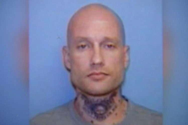 A close-up image of a man with a shaved head and prominent facial tattoos, standing against a blue background. He has a neutral expression and is wearing a gray shirt.