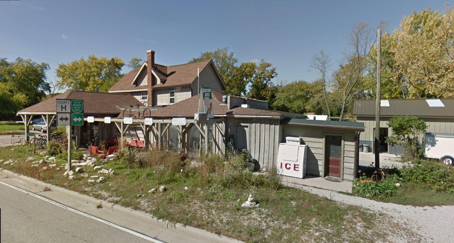 A quaint roadside building featuring a wooden structure with a porch, surrounded by greenery and rocks. Signs indicate directions and nearby attractions, while a vintage ice machine is visible on the side. The background includes trees and another building with a metal roof.