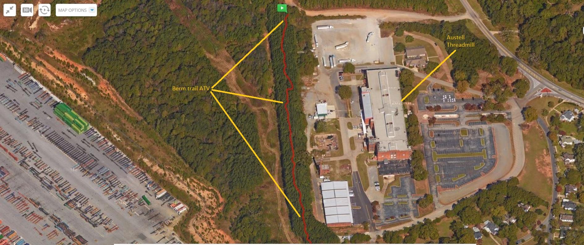 Aerial view of a wooded area with a marked berm trail for ATV use and a nearby industrial complex. The industrial area features a large building, parking lots, and multiple trucks. Labels indicate key locations, including "Berm trail ATV" and "Austell Threadmill." The image includes surrounding greenery and a road leading to the complex. Austell Threadmill MTB Park (Closed) mountain bike trail.