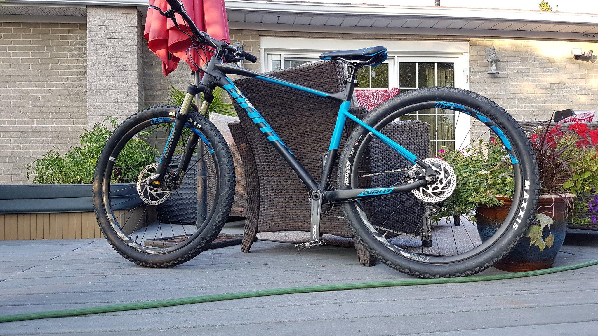 Giant Giant Fathom: A blue and black mountain bike leaning on a wooden deck, surrounded by potted plants and furniture. The bike features wide tires, a visible disc brake, and the brand name "Giant" prominently displayed on the frame. In the background, there are patio chairs and a reddish umbrella.