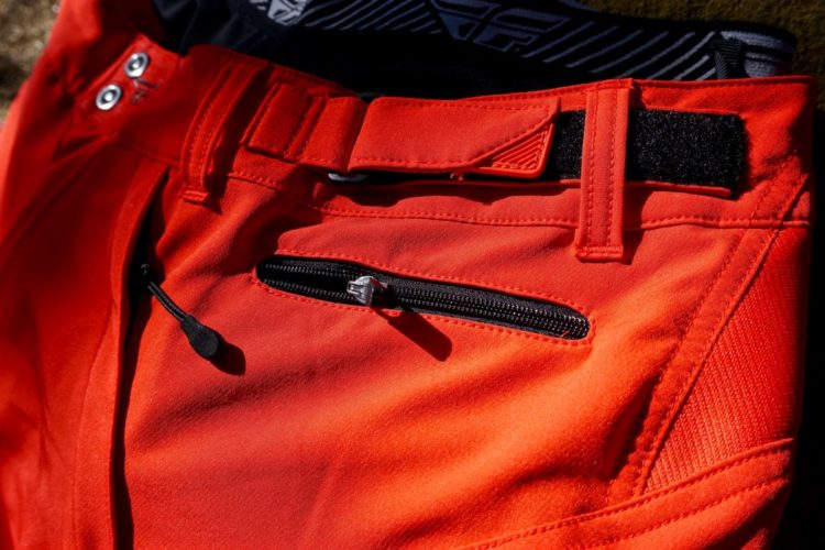 A close-up image of a pair of bright red pants, featuring a zippered pocket on the front and a sturdy waistband with a hook-and-loop closure. The fabric appears lightweight and durable, ideal for outdoor activities.