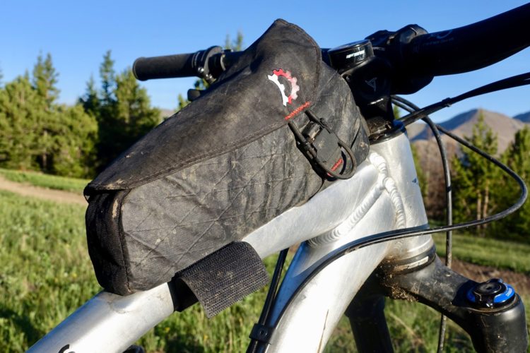 A close-up view of a bicycle's frame featuring a black top tube bag secured with straps. The background shows a green landscape with trees and a clear blue sky, suggesting an outdoor setting for cycling.