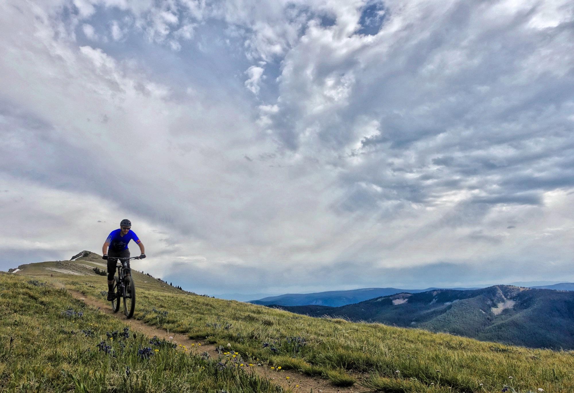 A mountain biker rides along a dirt trail on a grassy hillside, surrounded by wildflowers under a cloudy sky with dramatic cloud formations. In the background, rolling mountains stretch into the distance. Monarch Crest Trail mountain bike trail.