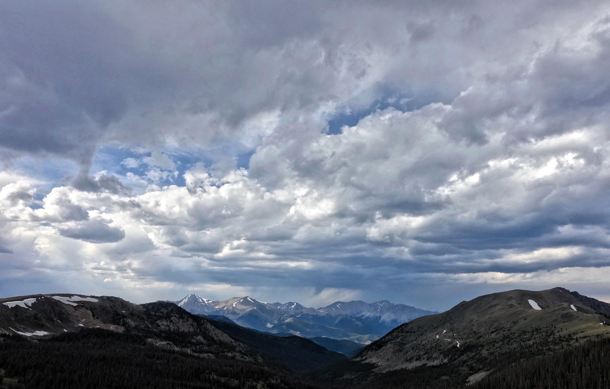 A panoramic view of a mountainous landscape under a dramatic sky filled with various shades of gray clouds. Snow-capped peaks rise in the background, while rolling hills and a dense forest are visible in the foreground. Sunlight breaks through the clouds, creating a striking contrast in the scenery. Monarch Crest Trail mountain bike trail.