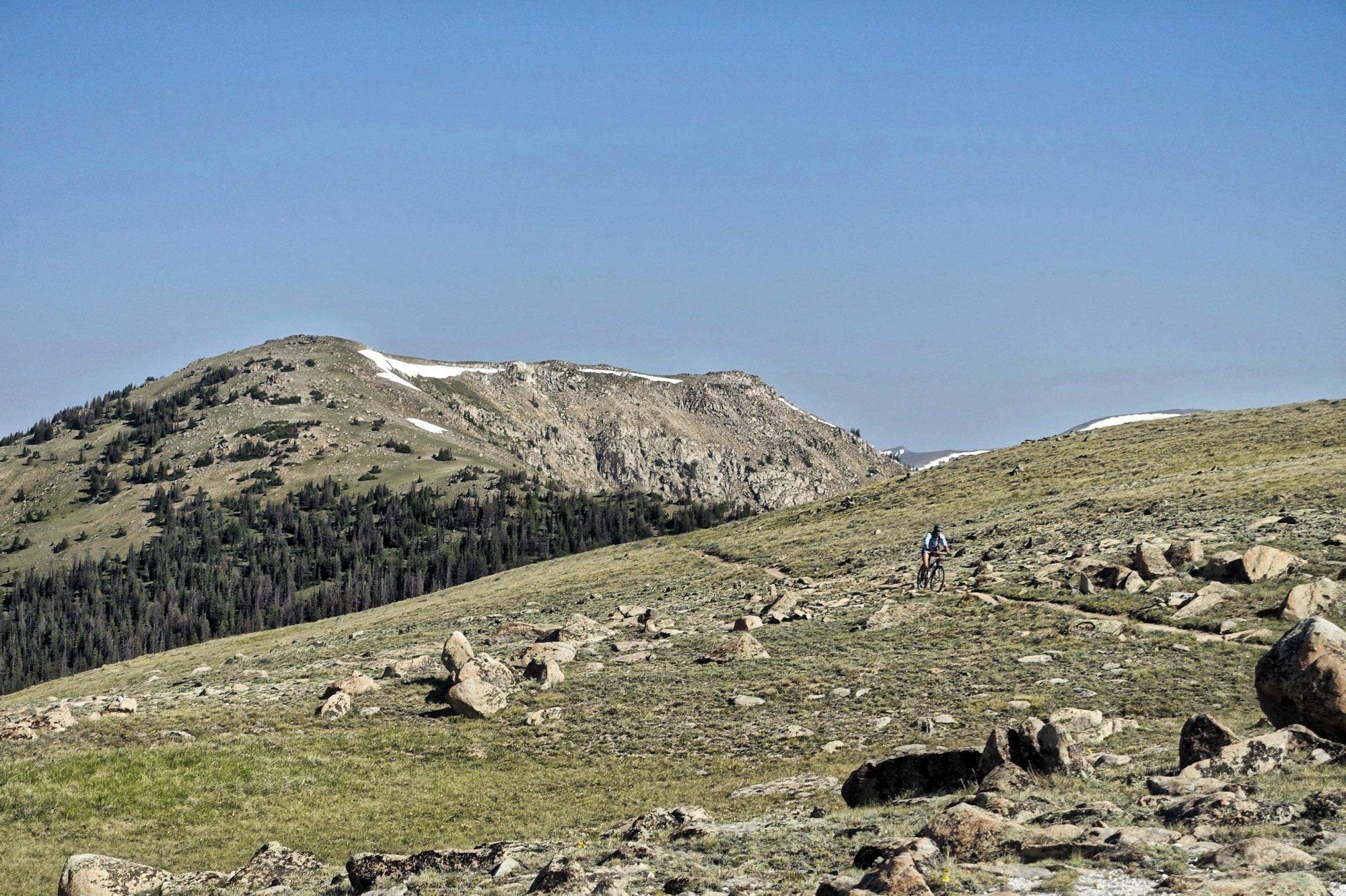 A mountain biker rides along a rocky trail on a grassy hillside, with lush green vegetation and distant snow-capped peaks under a clear blue sky. Monarch Crest Trail mountain bike trail.
