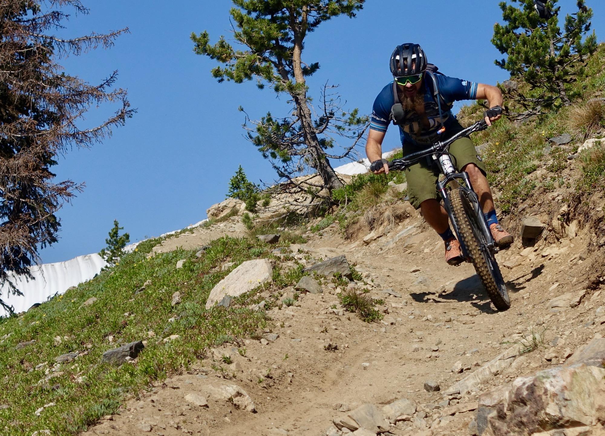 A mountain biker navigating a rocky trail surrounded by trees on a sunny day. The biker is leaning forward, focused on the terrain, with a clear blue sky in the background. Monarch Crest Trail mountain bike trail.