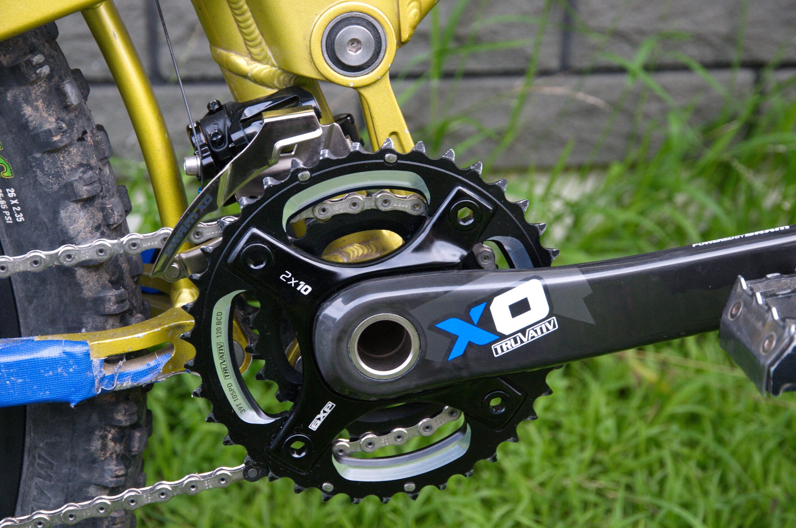 Mongoose Teocali Comp: Close-up view of a mountain bike's drivetrain, showcasing a black and silver crankset with dual chainrings, alongside a chain and a derailleur. The bike frame is a bright yellow color, and there is green grass in the background, with hints of a textured tire visible.