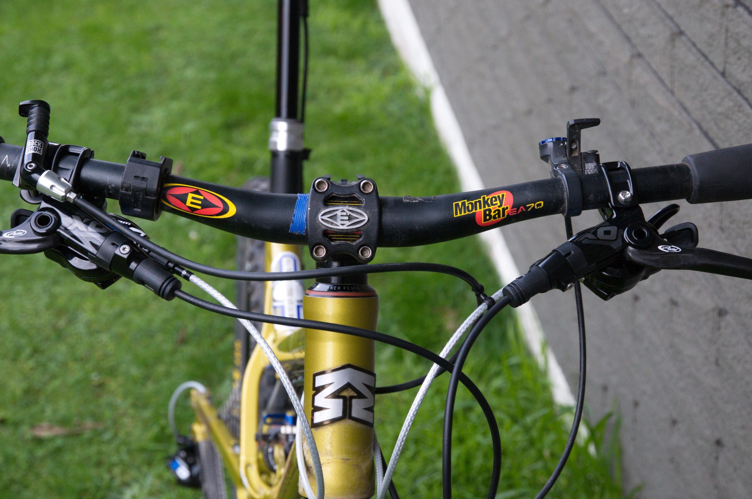Mongoose Teocali Comp: Close-up view of a mountain bike's handlebars, featuring black grips, a stem with colorful branding, and brake levers. The bike is positioned on grass with a grey wall in the background.