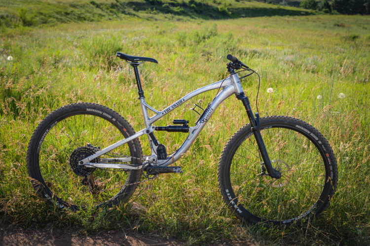 A mountain bike with a silver frame is positioned on grassy terrain, surrounded by tall grass and wildflowers. The bike features thick tires, a suspension system, and a sleek design, indicating it's built for off-road cycling.