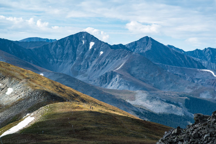 A scenic view of rugged mountain peaks under a partly cloudy sky, with grassy slopes in the foreground and patches of snow on the mountains, showcasing the natural beauty of a mountainous landscape with two hikers visible in the distance.
