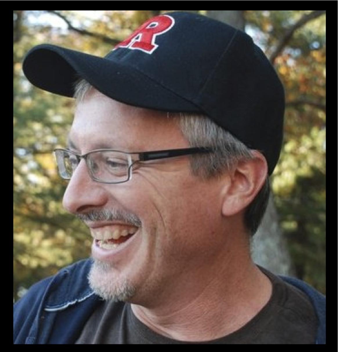 Cannondale Trail 6: A man with glasses and a black cap featuring a red letter R, smiling and laughing outdoors, with greenery in the background.