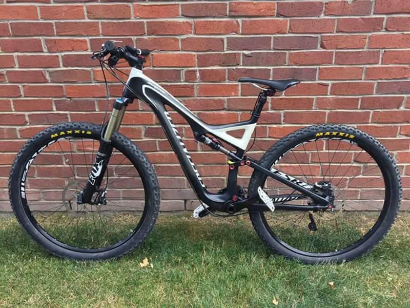 Specialized Stumpjumper FSR Comp Carbon 29: A black and white mountain bike stands against a red brick wall. The bike features thick tires with Maxxis branding, a sturdy frame, and a front suspension fork. It is positioned on green grass, highlighting its rugged design suitable for off-road riding.