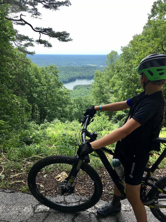 A young person wearing a helmet and cycling attire is standing beside a mountain bike on a trail, gazing out over a lush green valley. The view includes a lake surrounded by hills and dense forest under a cloudy sky. Lake Russell Loop mountain bike trail.