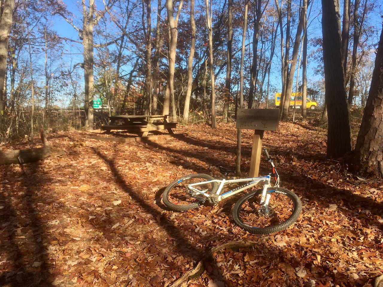 A white mountain bike resting on a carpet of autumn leaves in a wooded area, with a wooden sign labeled "HALL" nearby. In the background, a picnic table is visible, along with a green trail sign and a yellow vehicle passing on a road through the trees. The scene is illuminated by sunlight filtering through the branches. Chicopee Woods mountain bike trail.