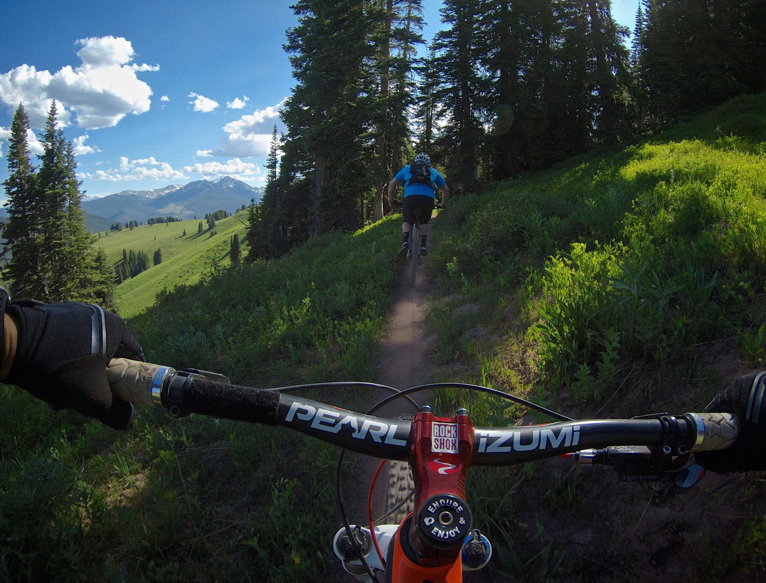 A mountain biker rides along a trail surrounded by lush greenery and tall trees, with mountains in the background under a blue sky with fluffy white clouds. The photo is taken from the perspective of another rider, showing the handlebars of the bike and a gloved hand gripping the handlebar. Vail Mountain Bike Park mountain bike trail.