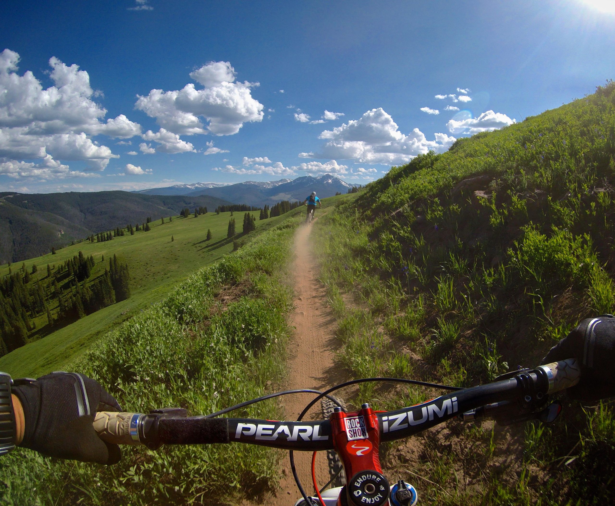 A mountain biker rides along a dirt path through lush green hills, with a panoramic view of distant mountains under a bright blue sky filled with clouds. The handlebars of another bicycle are visible in the foreground, emphasizing the biking perspective of the scene. Vail Mountain Bike Park mountain bike trail.