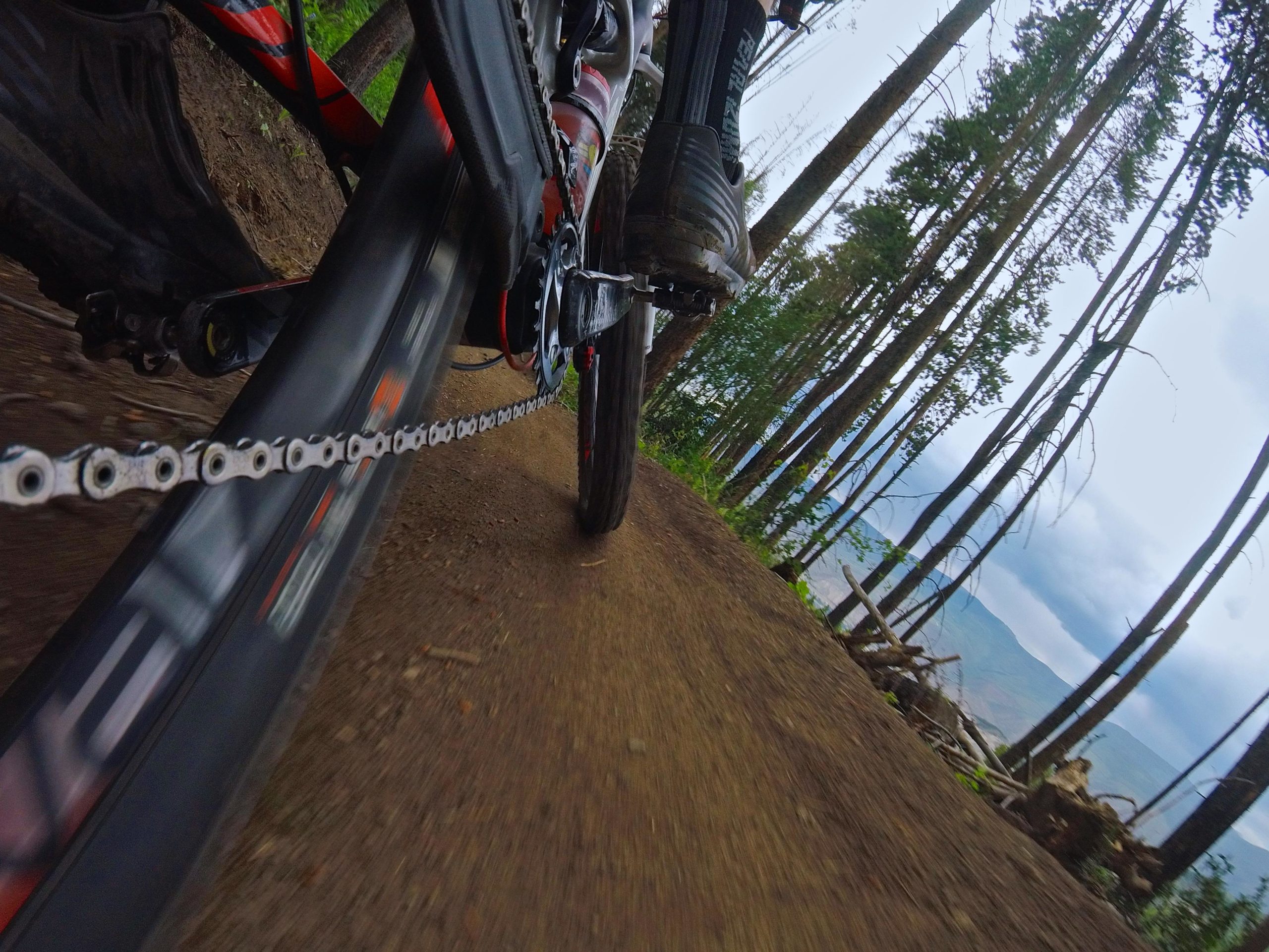 A close-up view of a mountain bike in motion along a dirt trail, featuring a focus on the bike's chain and tire. Surrounding the path are tall trees and a glimpse of a lake in the background under a cloudy sky. Beaver Creek Ski Resort mountain bike trail.
