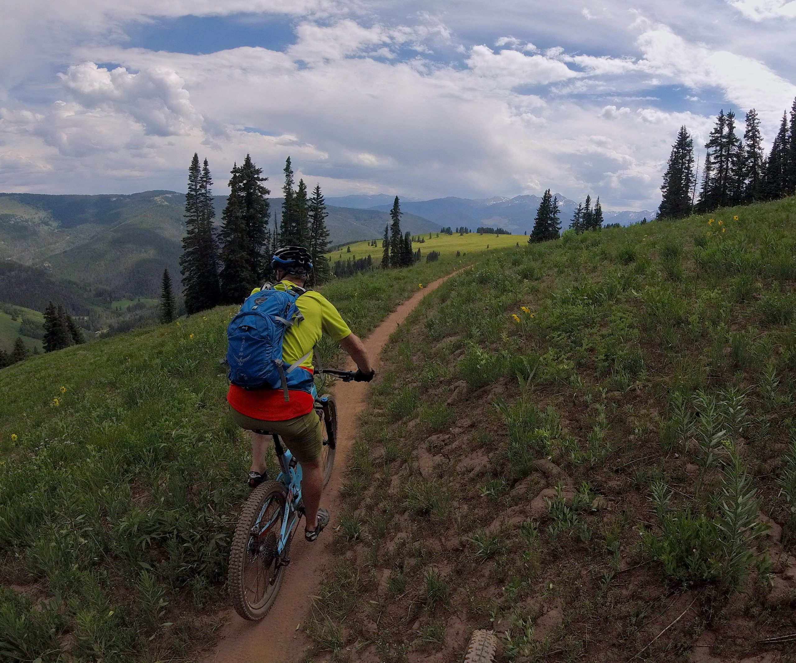 A person cycling along a dirt trail surrounded by lush green hills and tall pine trees, with a mountainous landscape in the background under a partly cloudy sky. The cyclist is wearing a bright yellow shirt and a blue backpack. Vail Mountain Bike Park mountain bike trail.