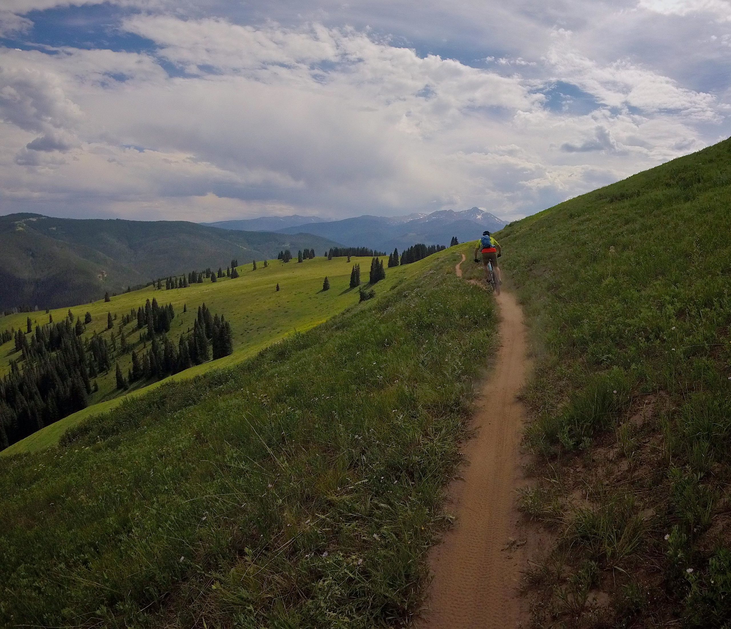 A cyclist riding along a winding dirt trail on a grassy hillside, surrounded by lush green meadows and distant mountains under a partly cloudy sky. Vail Mountain Bike Park mountain bike trail.