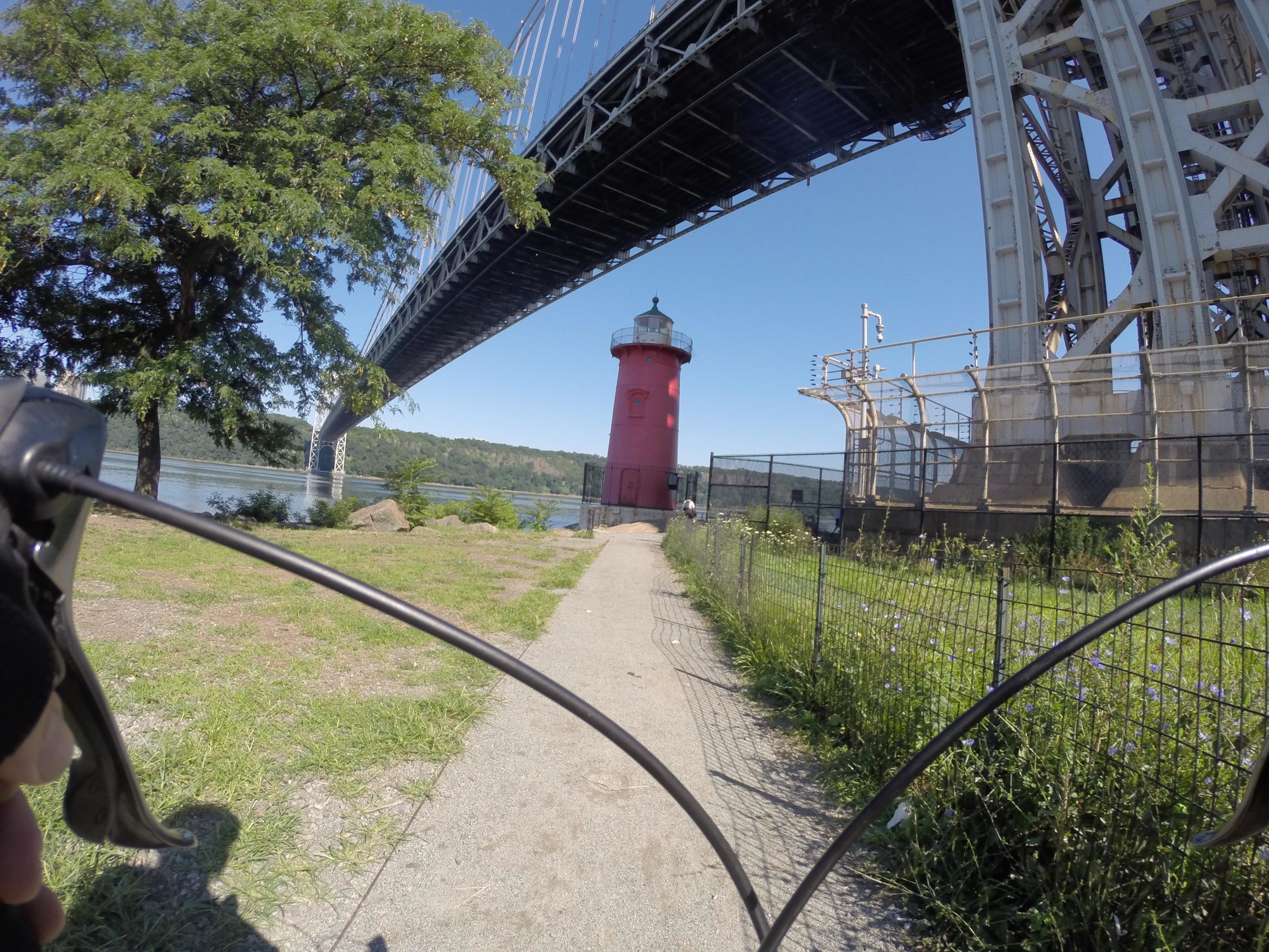 A view from a bicycle perspective along a path leading to a red lighthouse situated near the water, with a large bridge above it. Lush greenery is visible on the left, and a fence runs alongside the path. The scene is set on a clear blue sky day. West Street Greenway mountain bike trail.