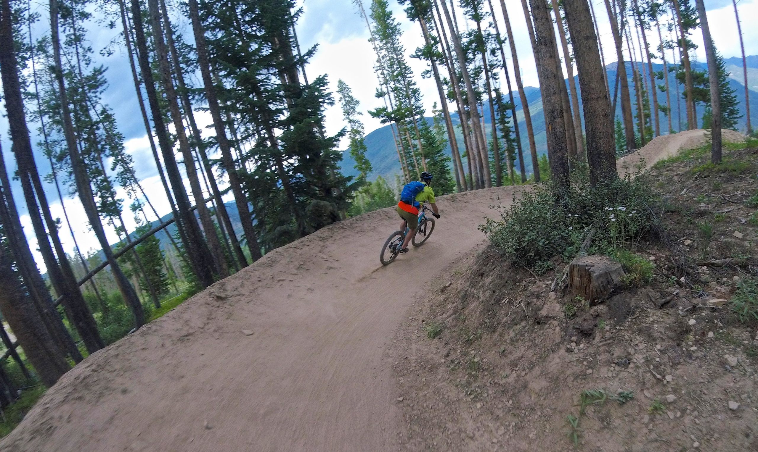 A mountain biker riding along a dirt trail through a forest of tall trees and distant mountains under a cloudy sky. The biker is wearing a bright green shirt, orange shorts, and a blue backpack, leaning into a turn on the rugged terrain. Vail Mountain Bike Park mountain bike trail.
