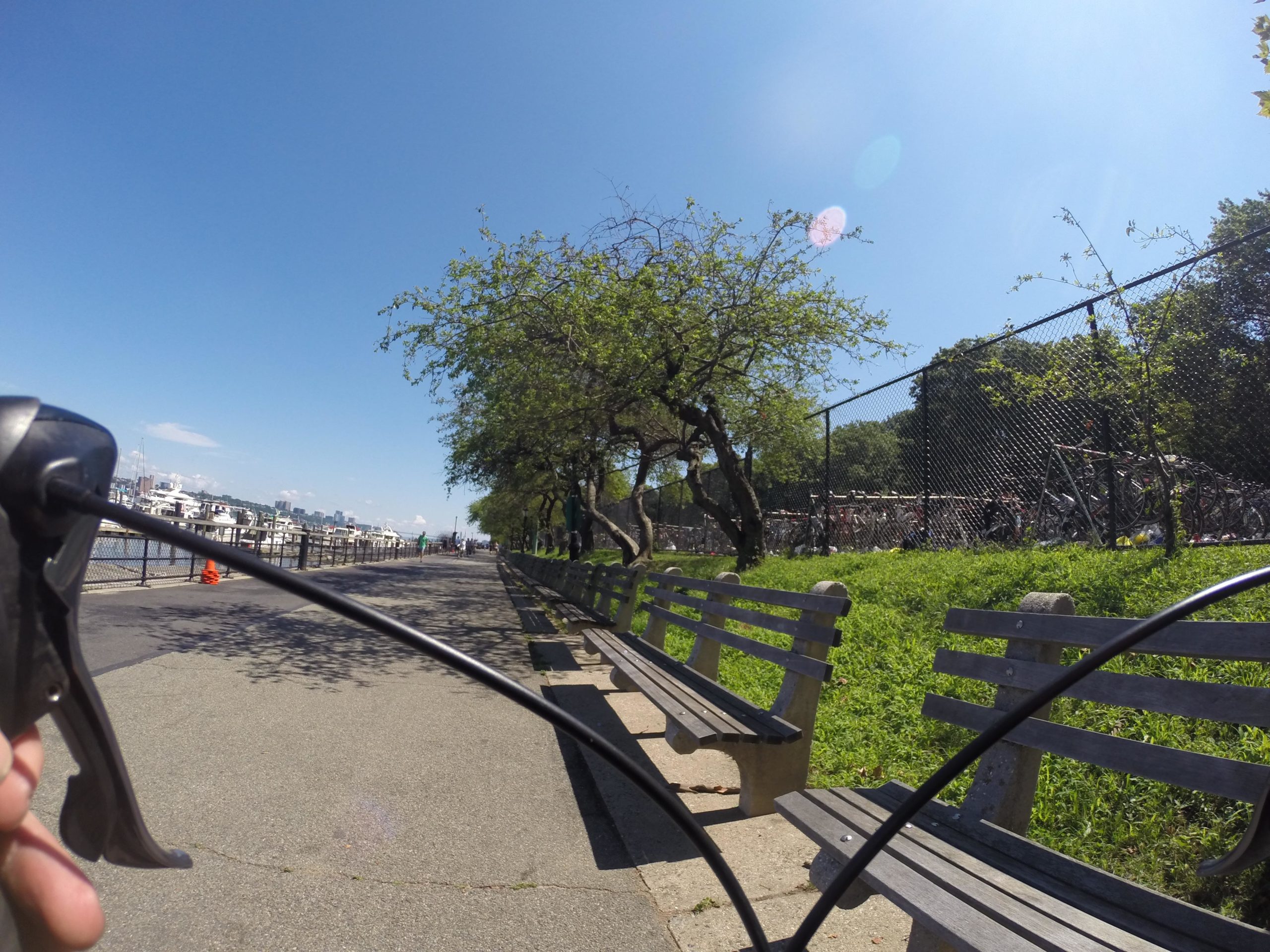 A scenic view of a waterfront pathway lined with benches and trees, overlooking a marina. The perspective shows a person's hand holding a bicycle handlebar, with shadows from trees on the pavement. In the background, there are parked bicycles behind a fence and a clear blue sky. West Street Greenway mountain bike trail.