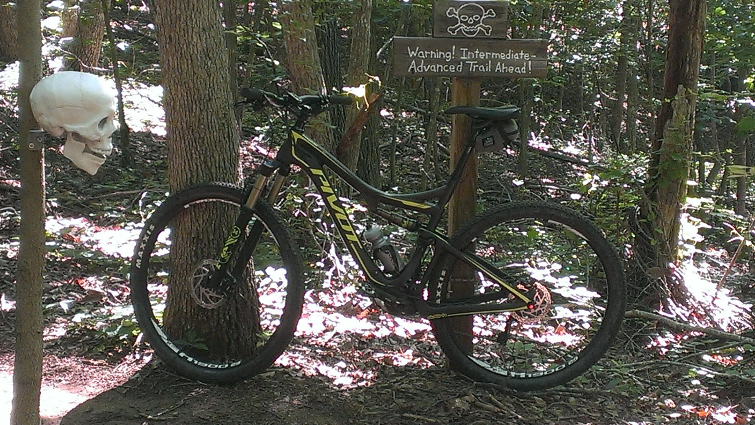 A mountain bike leaning against a tree near a sign that warns of an intermediate to advanced trail ahead. A decorative skull is mounted on a nearby tree, adding an adventurous flair to the forest setting. Sunlight filters through the leaves, highlighting the bike and the natural surroundings. Chicopee Woods mountain bike trail.
