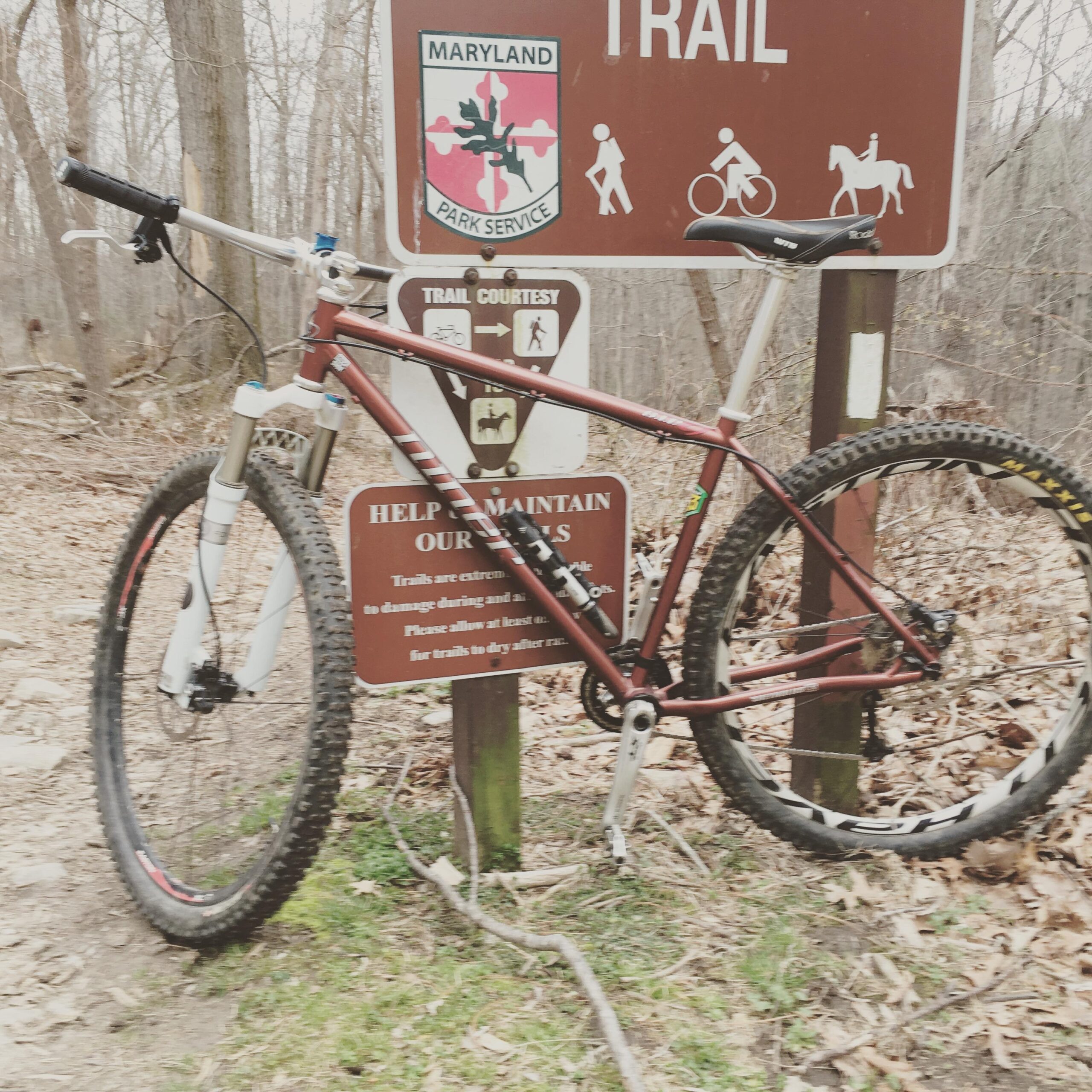 Niner S.I.R. 9: A mountain bike resting against a trail sign in a wooded area. The sign features information about trail use and courtesy, with symbols indicating allowed activities for hikers, cyclists, and horseback riders. The surrounding environment is sparse with fallen leaves, and bare trees suggest early spring or late fall.