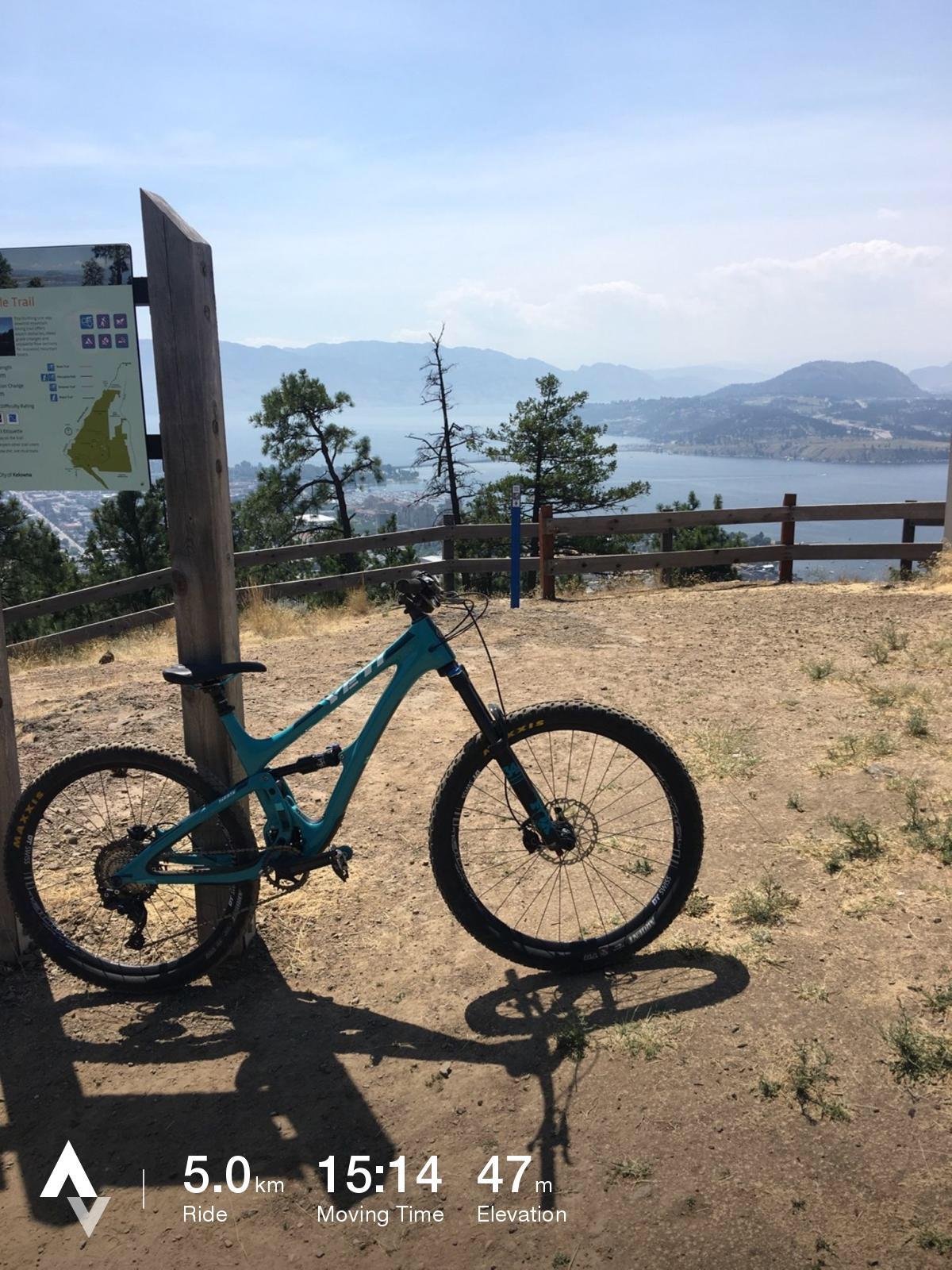 Yeti SB5: A mountain bike parked next to a trail sign with scenic views of a lake and mountains in the background. The image displays the bike's shadow on the ground, and the trail information indicates a ride distance of 5.0 km, a moving time of 15:14 minutes, and an elevation gain of 47 meters.